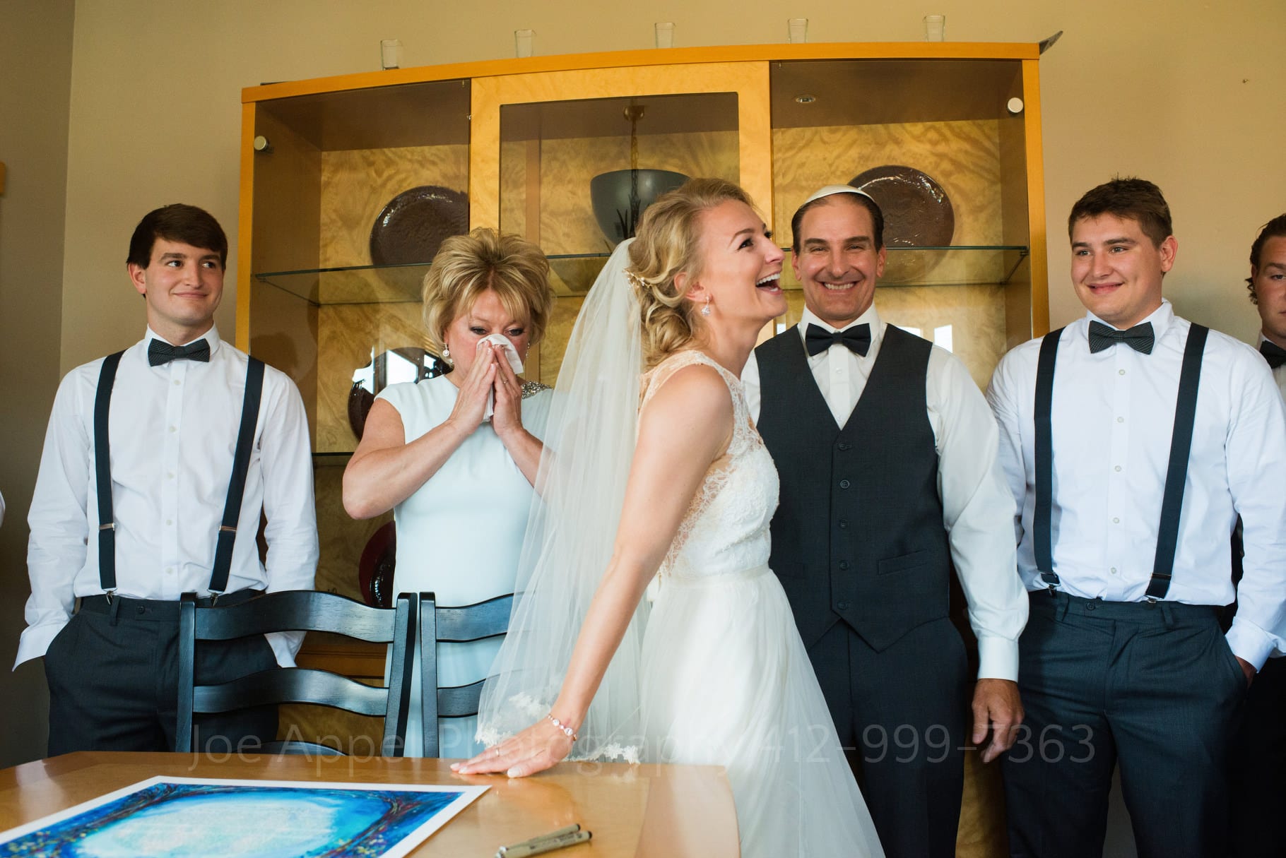 A bride smiles as she rises from a table after signing a blue-colored ketubah. Her mother wipes tears from her eyes while other witnesses smile.