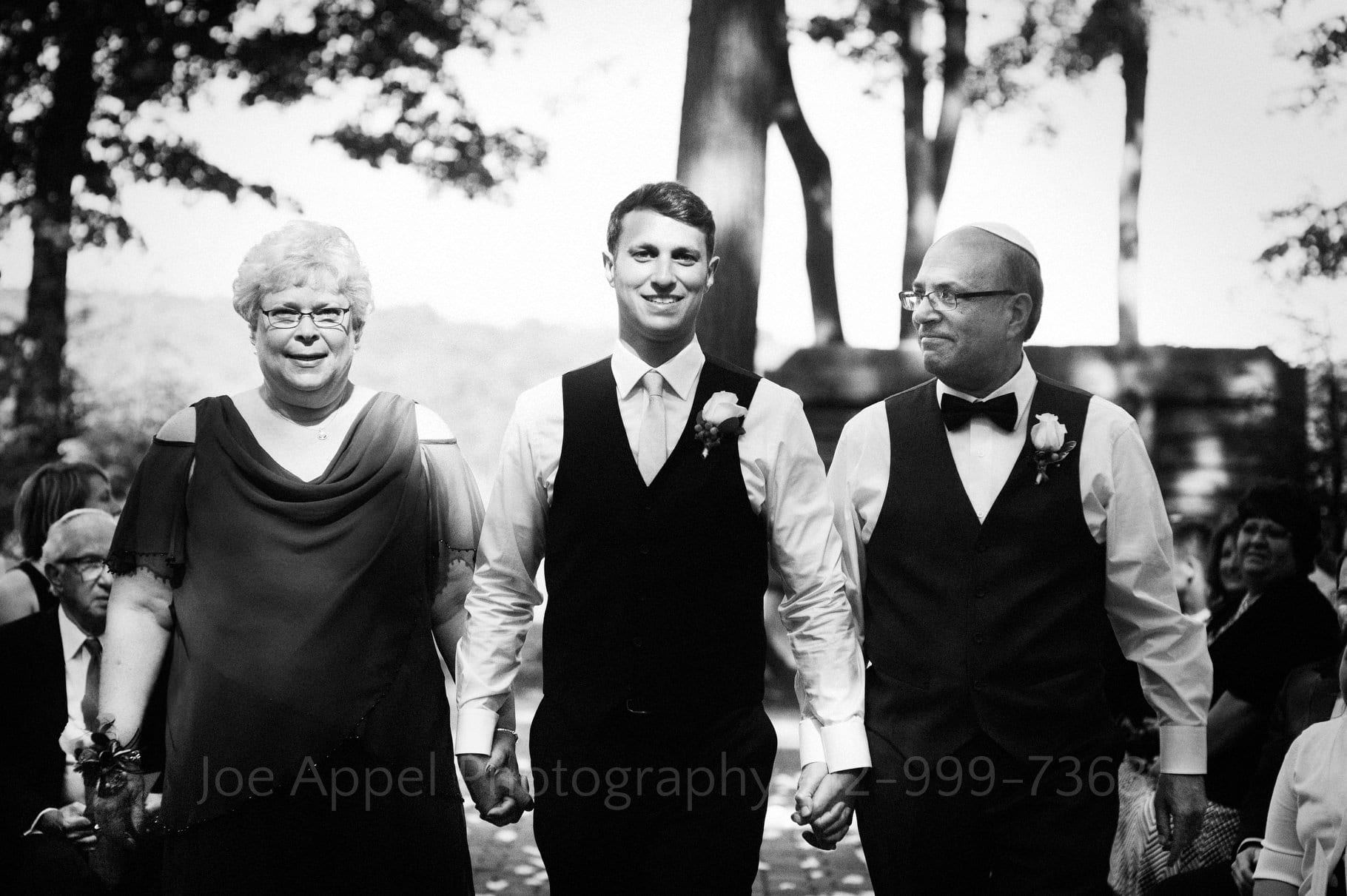 A groom holds hands with his mother and father as he walks down the aisle in a forest grove.