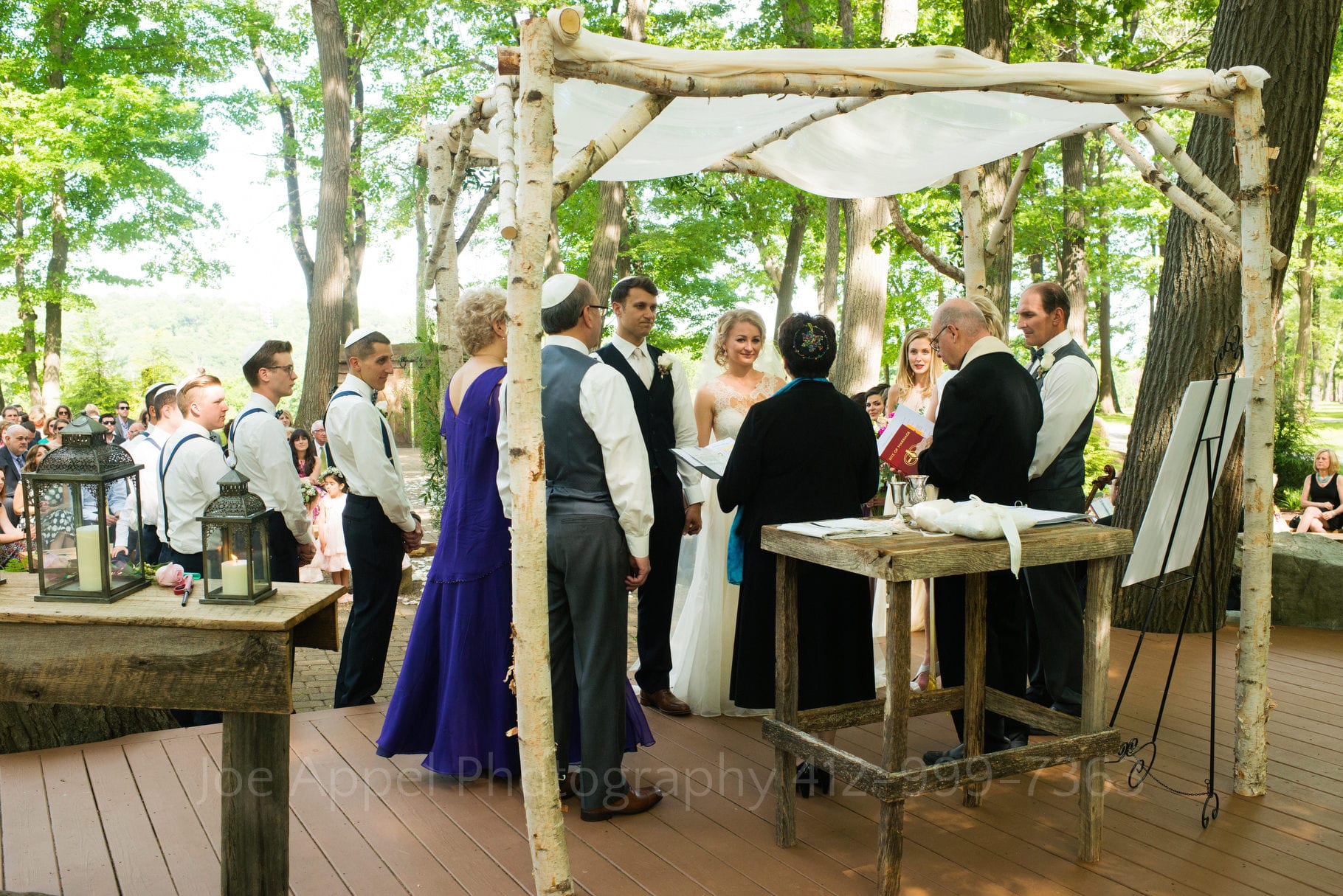 Parents of the couple surround them as they all stand beneath a chuppah set up in a forest grove for an outdoor wedding ceremony.