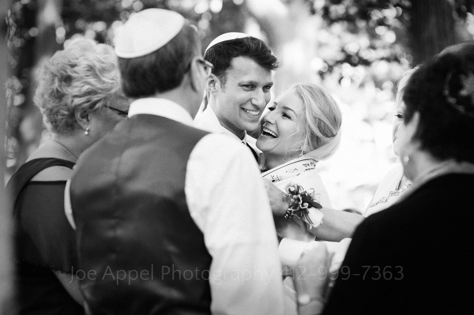 A bride and groom smile as they are wrapped together in a prayer shawl during their Seven Springs Wedding.