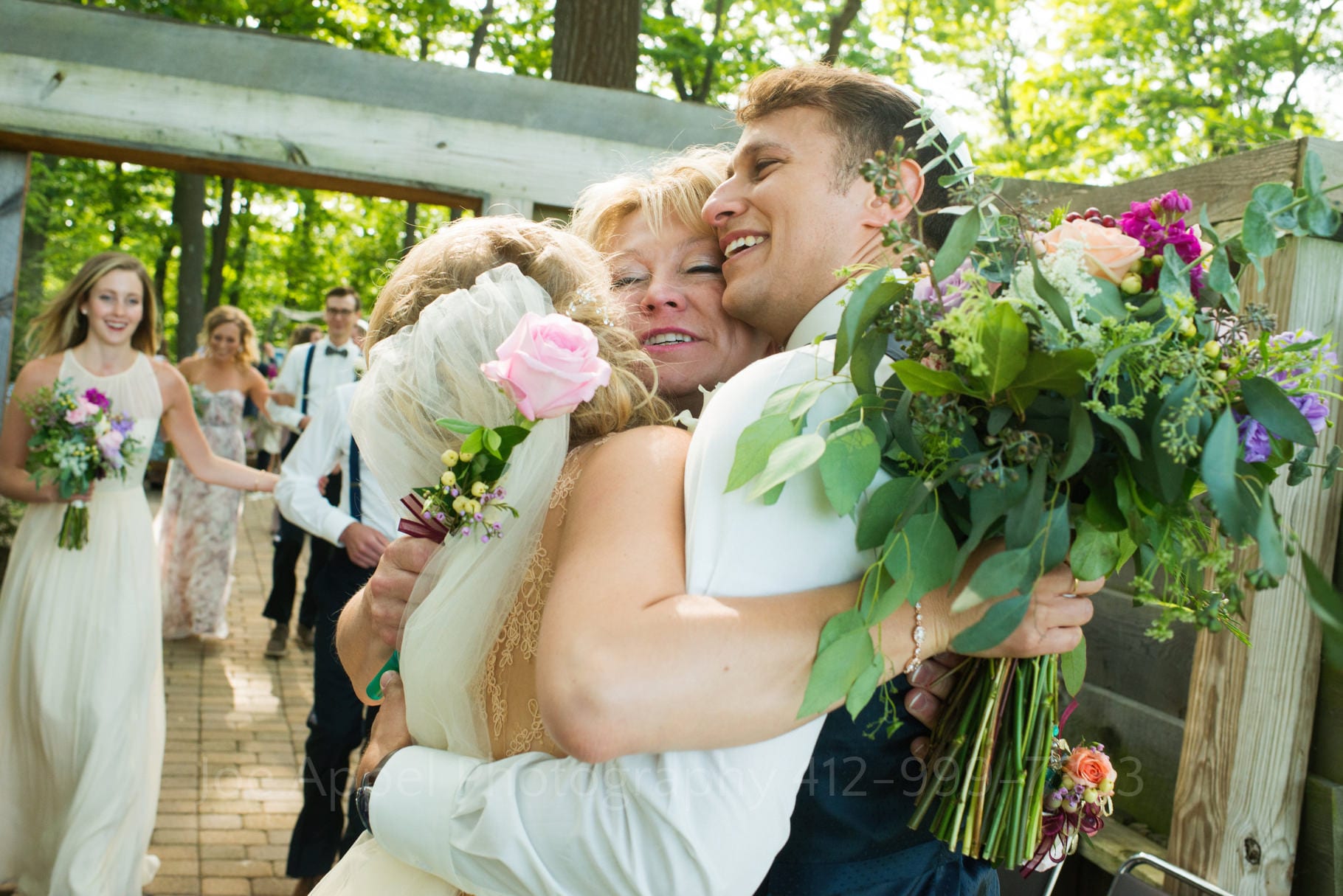A bride and groom smile as they are embraced by the bride's mother during their Seven Springs Wedding.