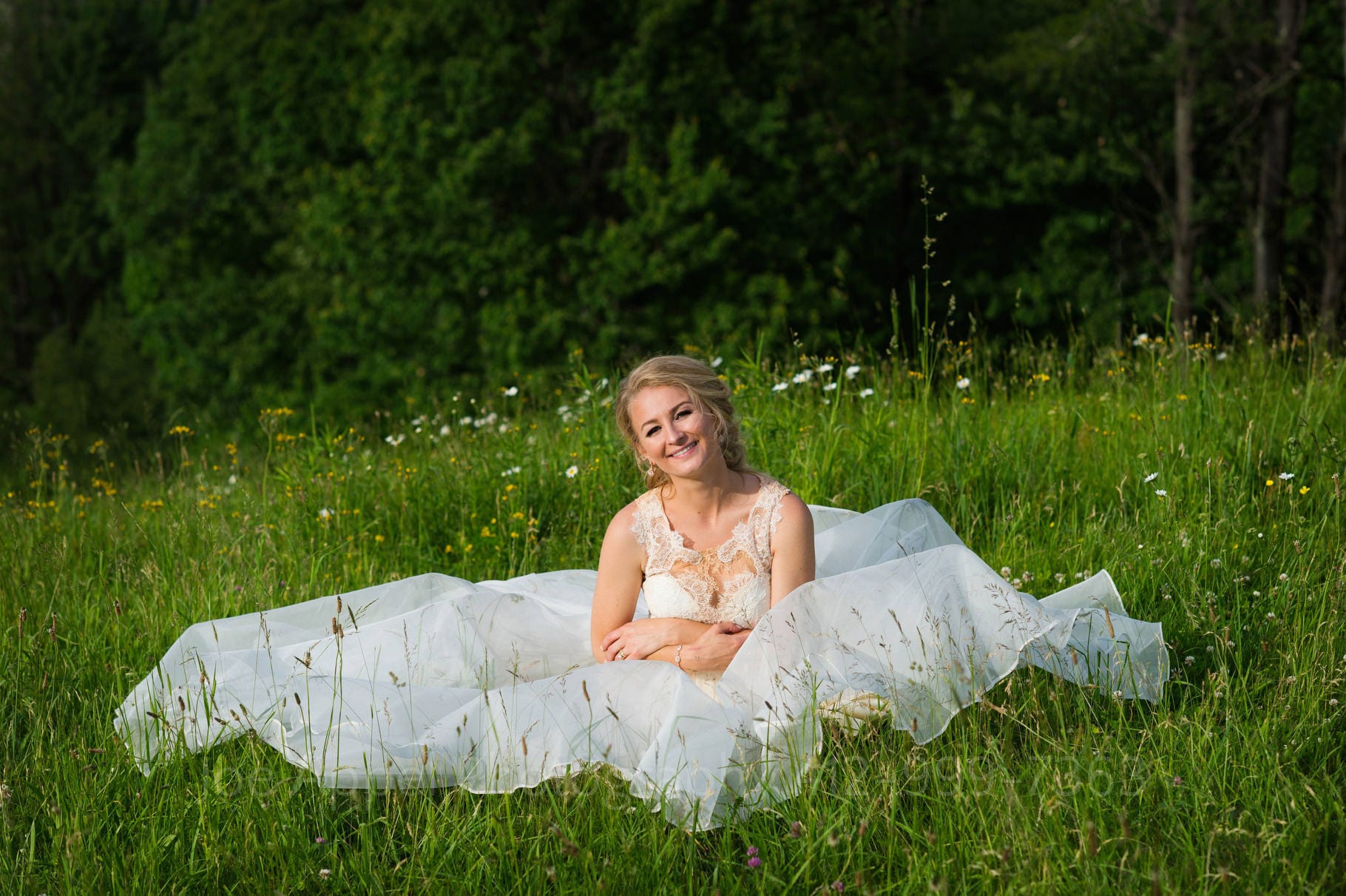 A bride smiles as she sits in tall grass in a mountain meadow at Seven Springs resort with her dress spread out around her during their Seven Springs Wedding.