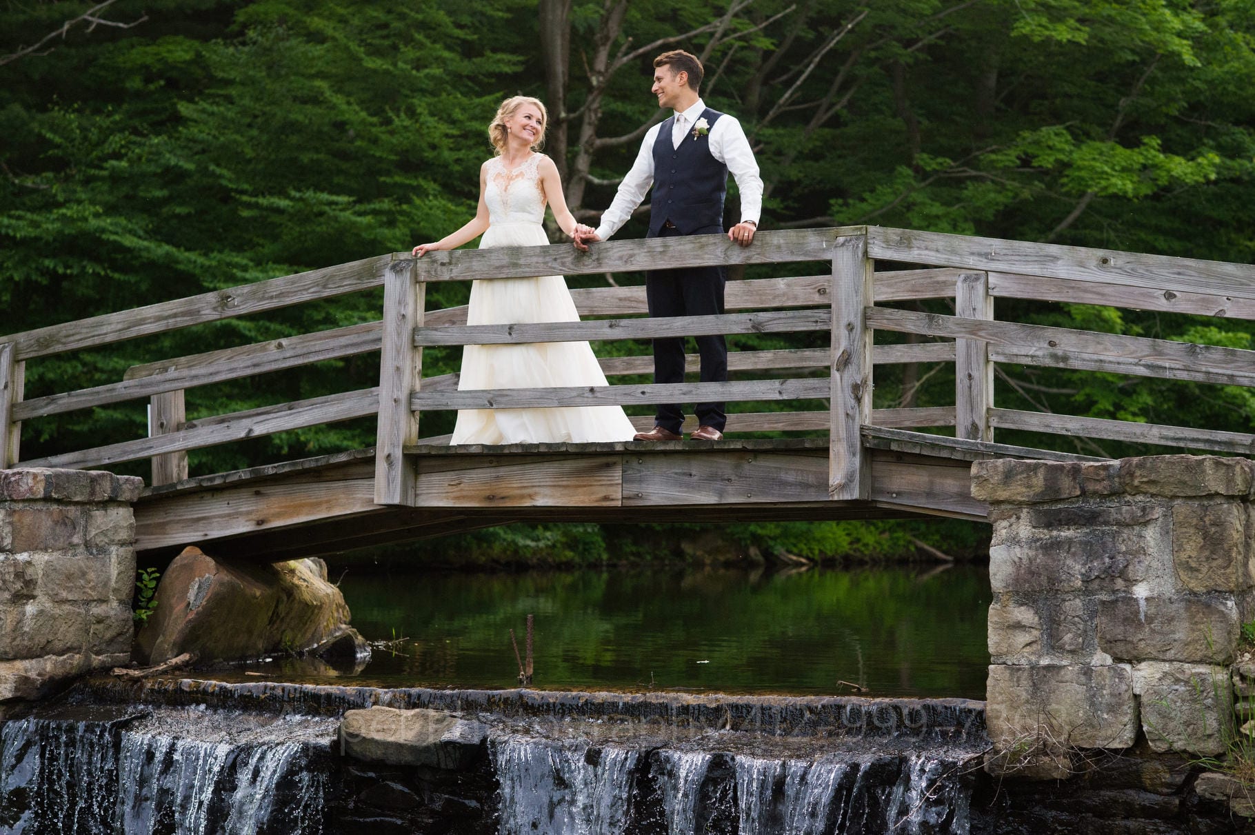 A bride and groom stand on a small wooden bridge above a dam at a pond during their Seven Springs Wedding.