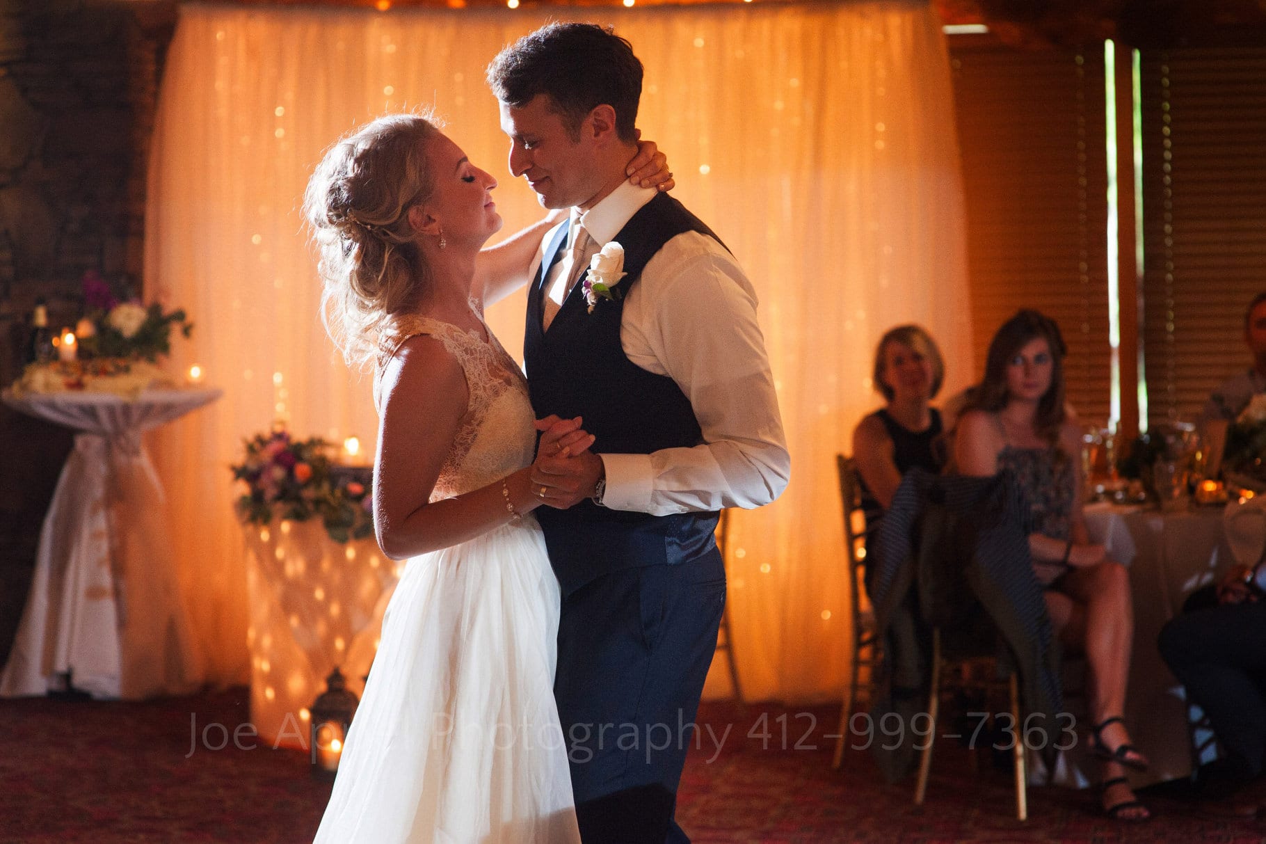 A bride and groom have their first dance in front of a warm, glowing curtain in a ballroom during their Seven Springs Wedding.