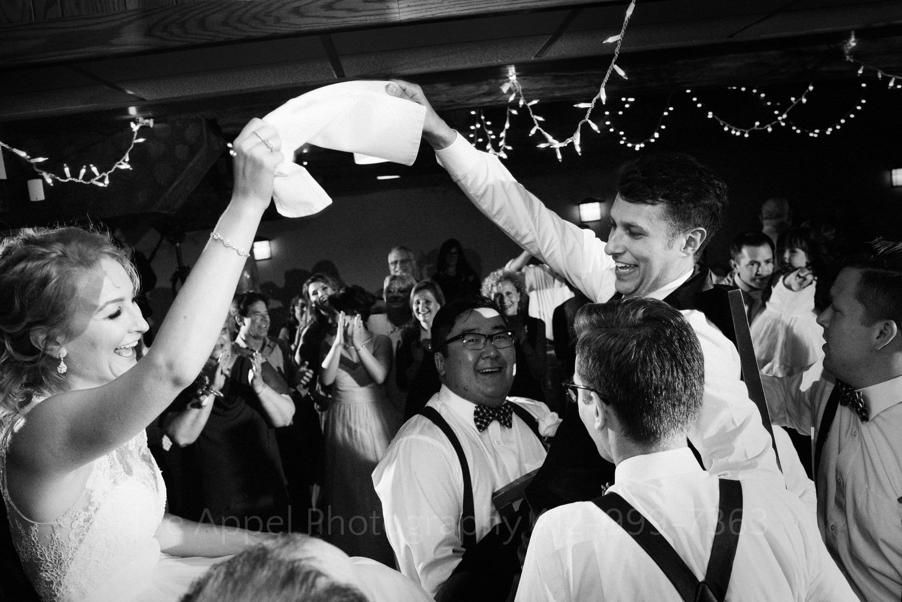 A bride and groom are lifted in chairs as they hold a napkin between them while dancing the Hora during during their Seven Springs Wedding.
