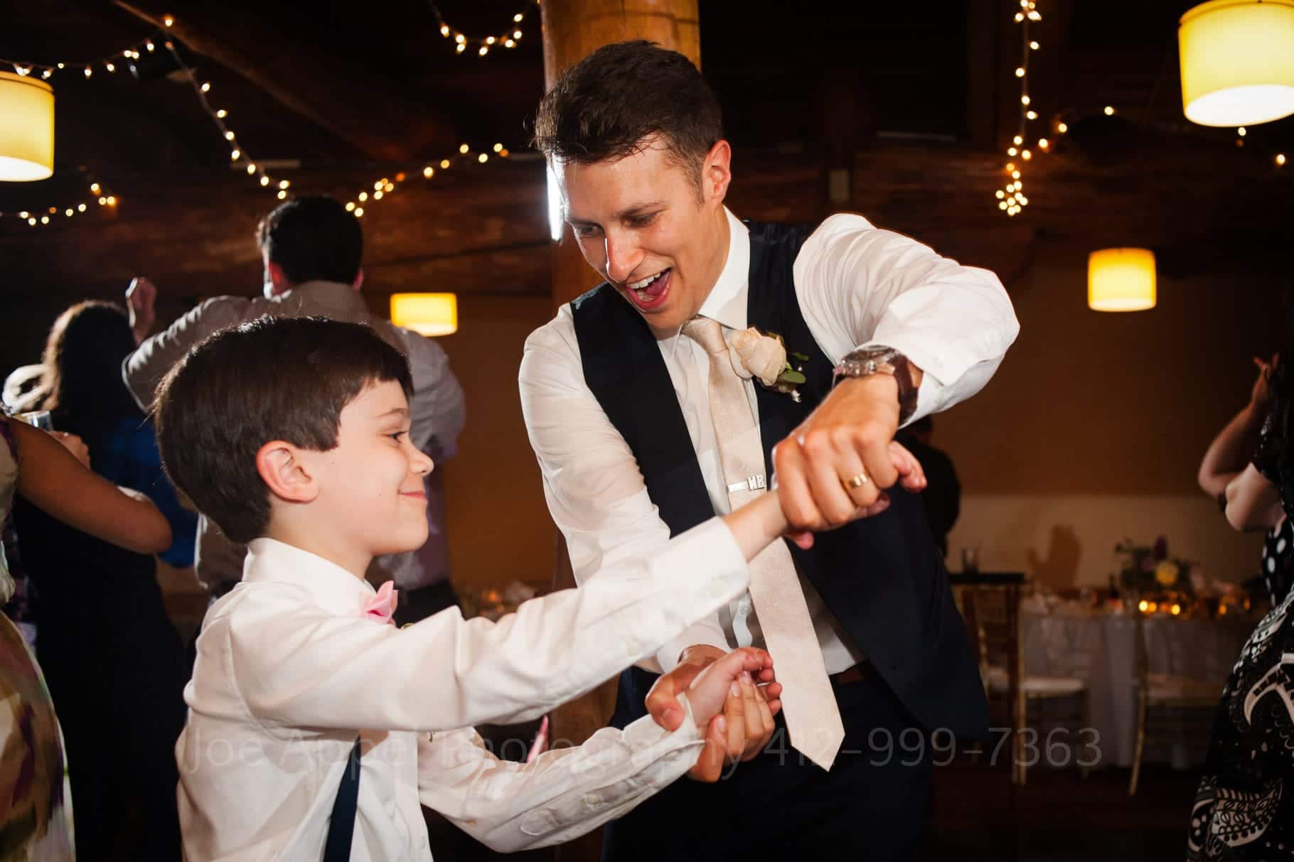 A groom in a vest and shirtsleeves dances with a young boy during during their Seven Springs Wedding.