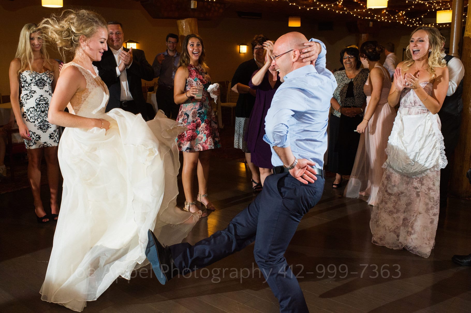 A bride kicks her feet up into the air as she dances with a guest in a blue shirt with one hand behind his head kicking his feet into the air during during their Seven Springs Wedding.