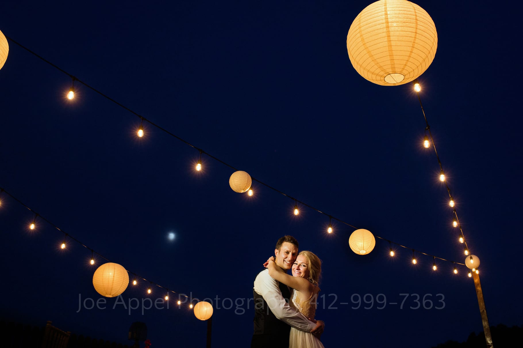 With chinese paper lanterns providing round dots of warm light against the twilight sky, a bride and groom embrace and smile at the camera on the deck outside of the Alpine Room during their Seven Springs Wedding.