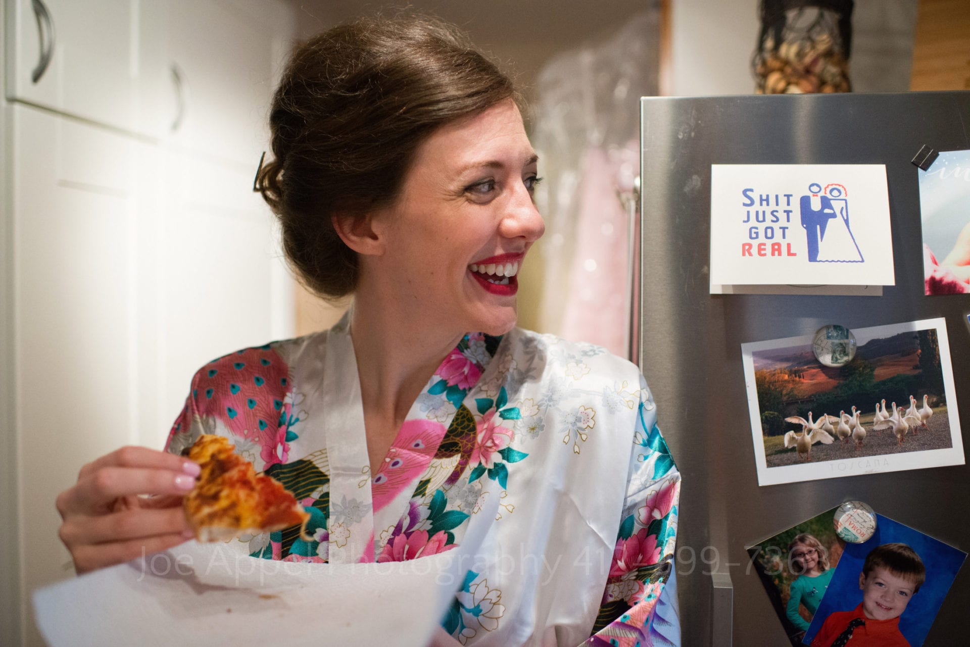 A bride wearing a floral dressing gown laughs as she eats a piece of pizza before her wedding. She's looking at a card that says "Shit just got real"