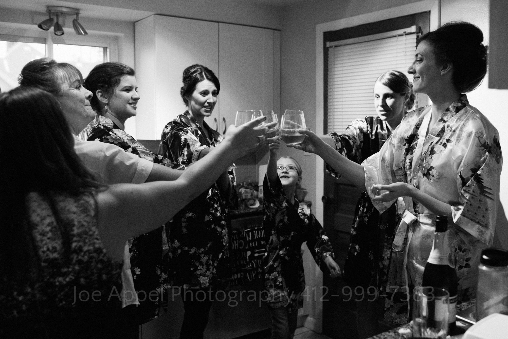 A bride and her bridesmaids touch glasses as they toast each other in a crowded kitchen.