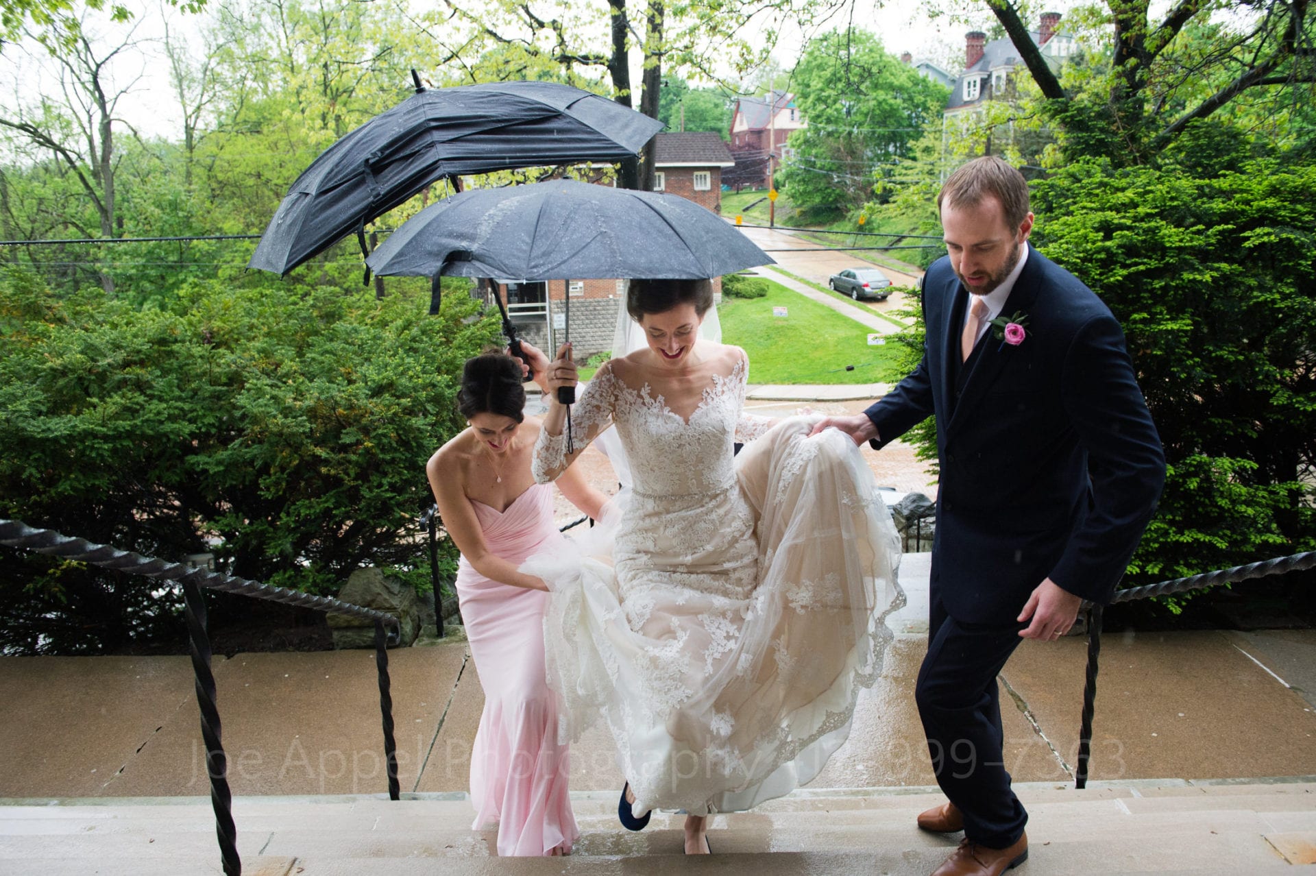 A bride is helped up the stairs leading to the church by her brother and sister. It's raining so they're holding umbrellas.