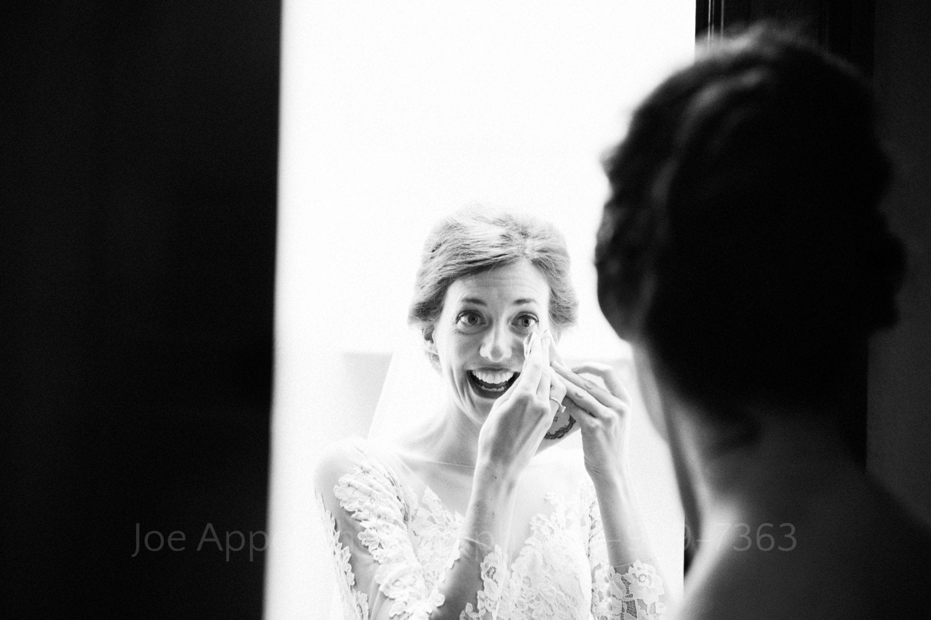 Black and white photo of a bride smiling as she wipes a tear from her eye as she stands in a room before the processional that begins her wedding.