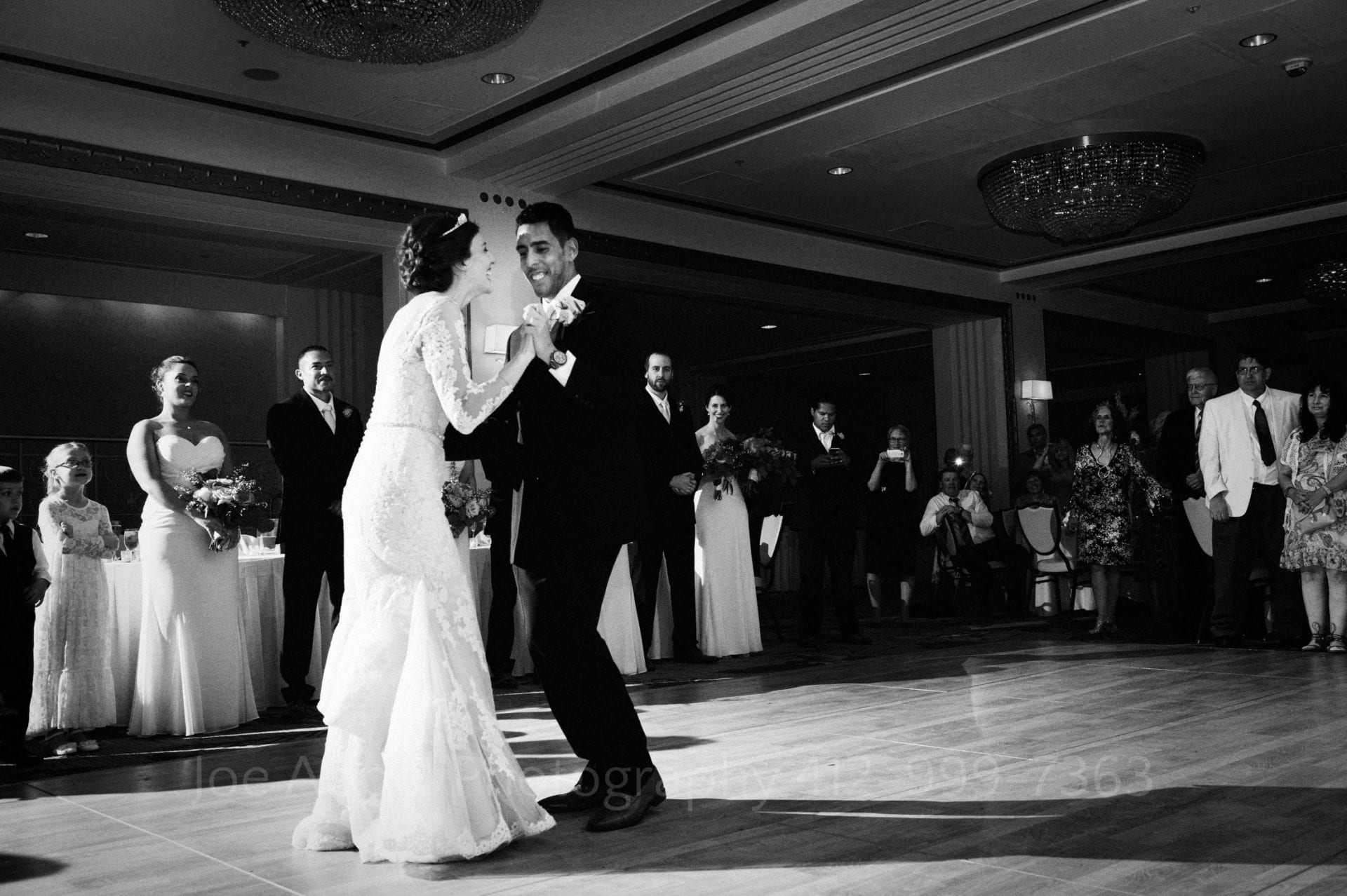 Bride and groom hold on to each other and laugh during their first dance at their William Penn Hotel Wedding in the William Penn ballroom as their wedding party stands to watch.