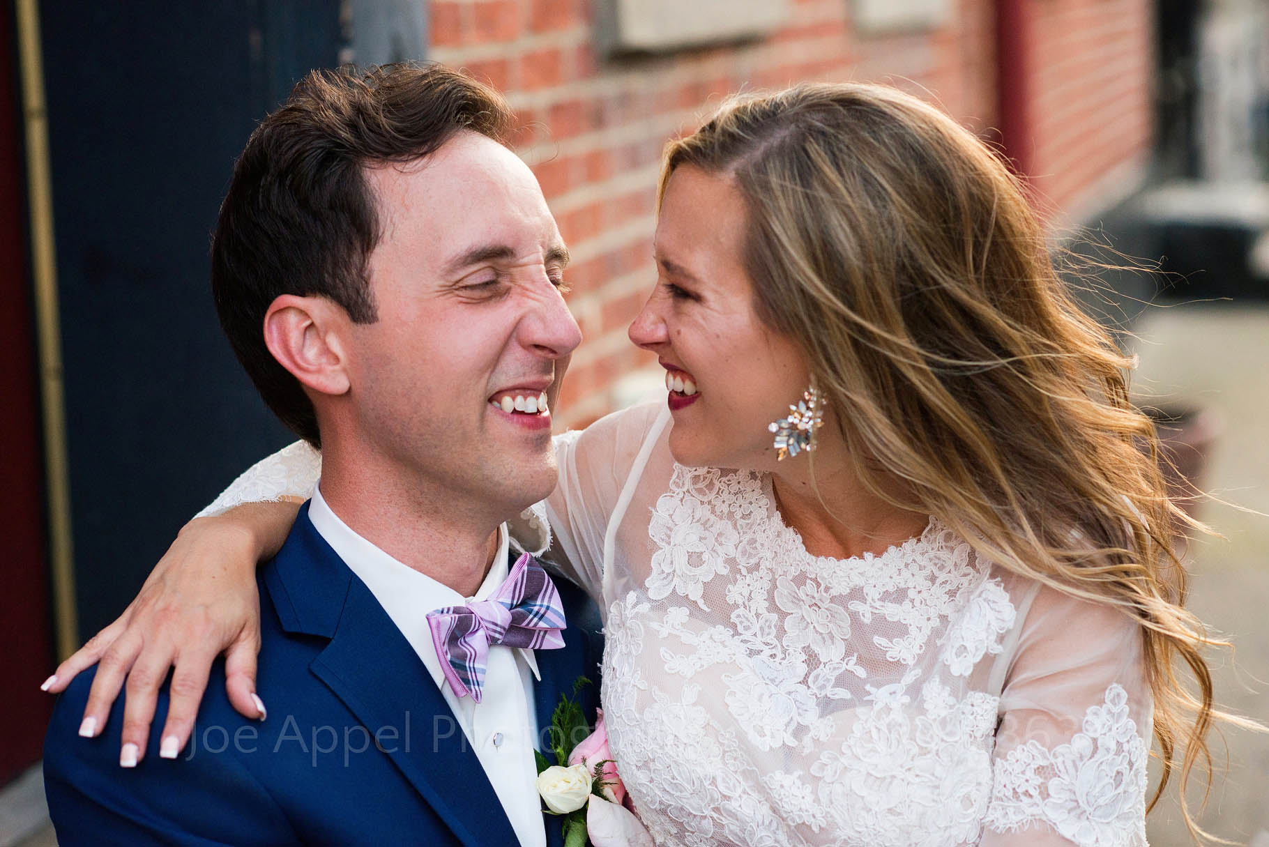 Young married couple look at each other and smile. Bride has her arm around the groom who is wearing a dark blue suit and a pink plaid bow tie.