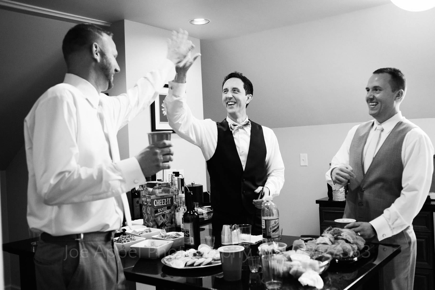 Three men have drinks around a table with booze and snacks on it. Two of the men are exchanging a high five while the third one laughs.