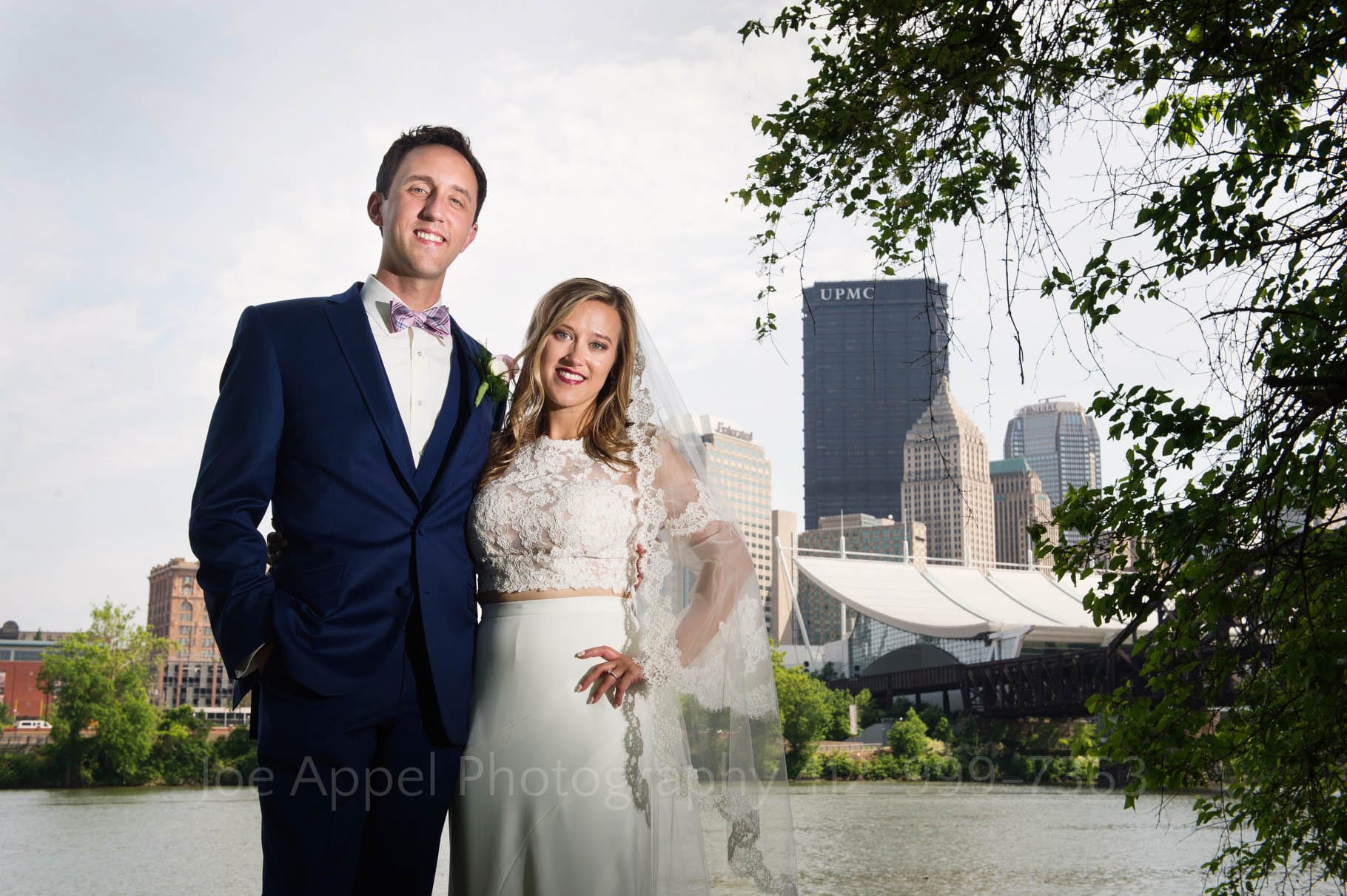 Portrait of a bride and groom as they stand together beneath a tree with the Allegheny River and a view of downtown Pittsburgh behind them.