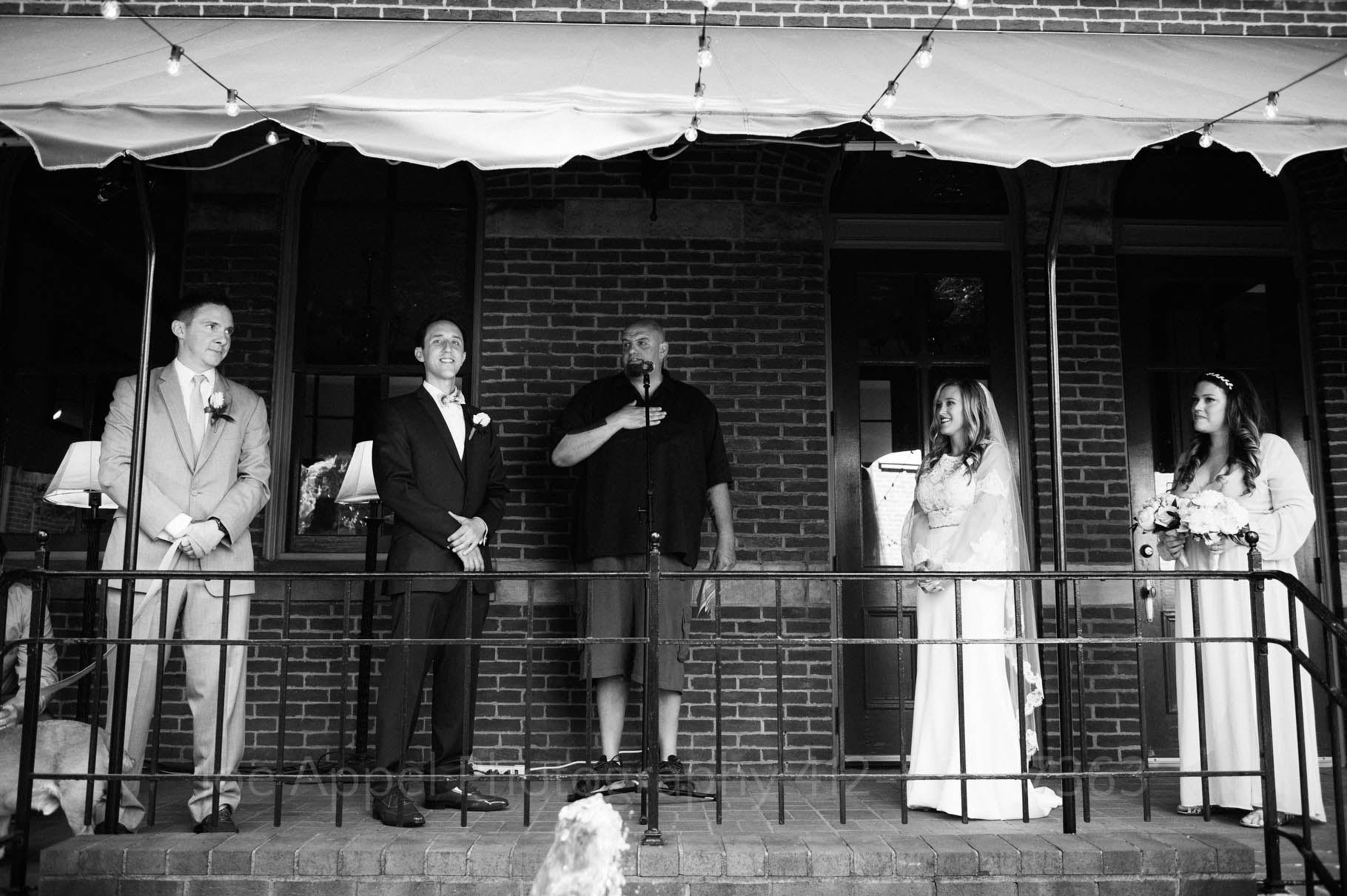 Braddock PA mayor John Fetterman addresses the audience gathered for a wedding. The bridal party stands on a porch beneath an awning.