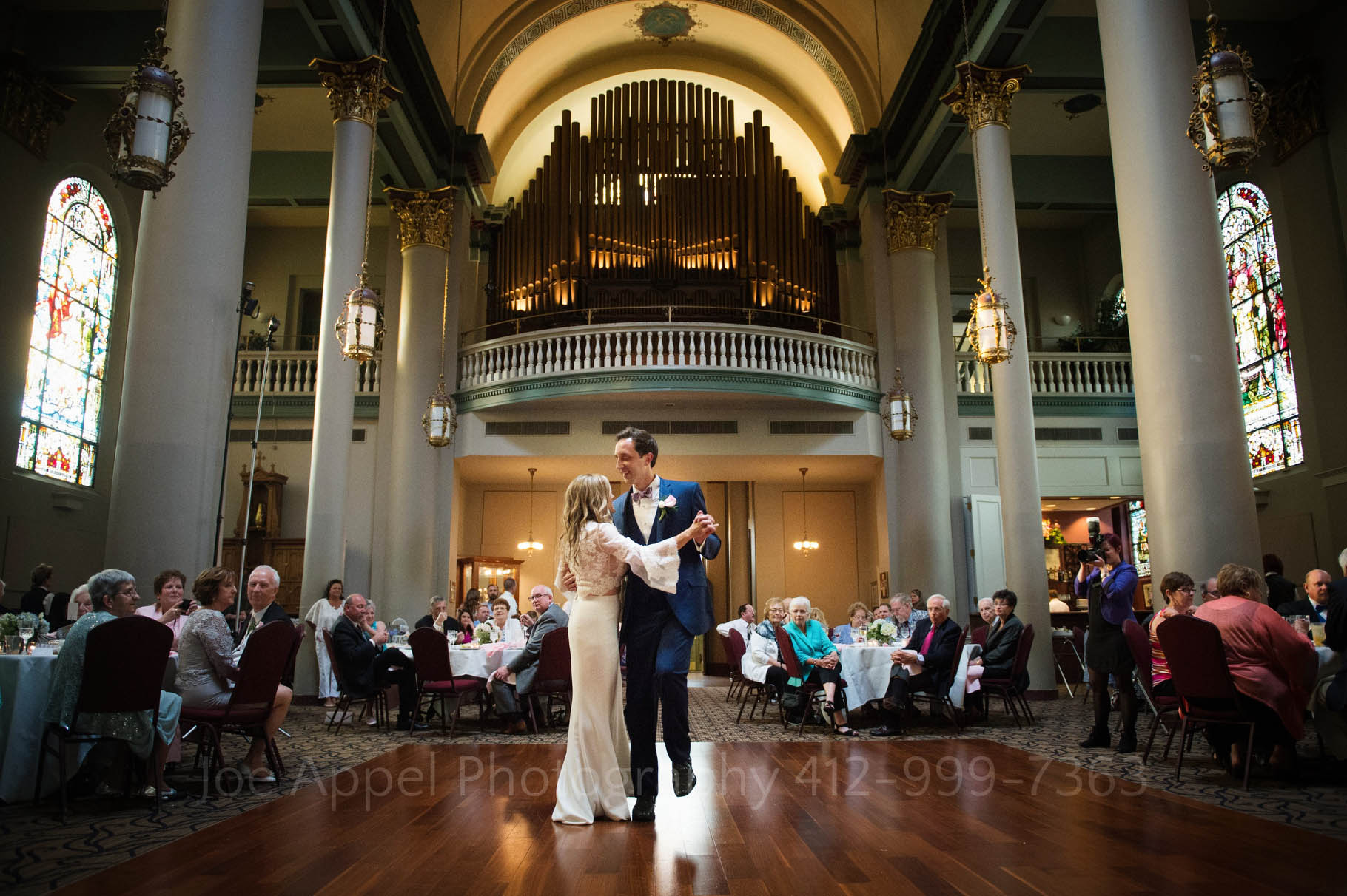 Groom raises his left foot into the air as he and his bride share their first dance during their Pittsburgh Grand Hall Wedding. The dance floor is in the center of the room with stained glass windows on either side and a large, backlit pipe organ in the loft above them.