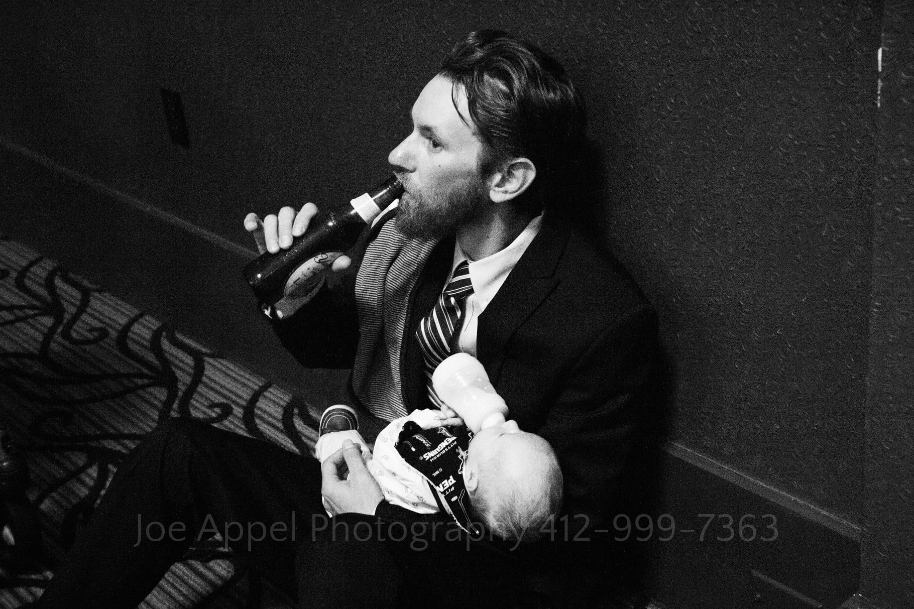 A man sits against a wall and sips a bottle of beer while holding his infant who drinks a bottle of milk.