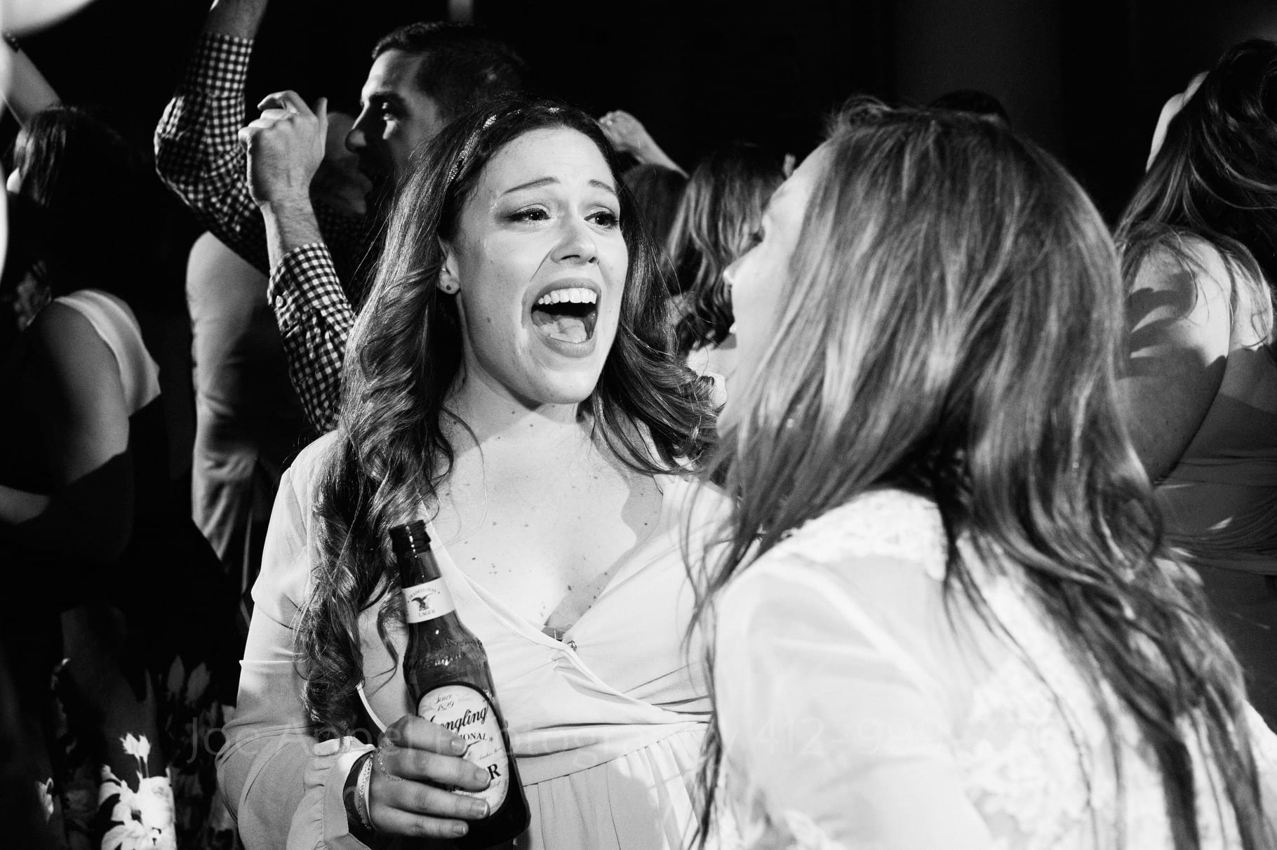 A bridesmaid holds a bottle of beer as she sings along with a bride on the dancefloor at her wedding reception.