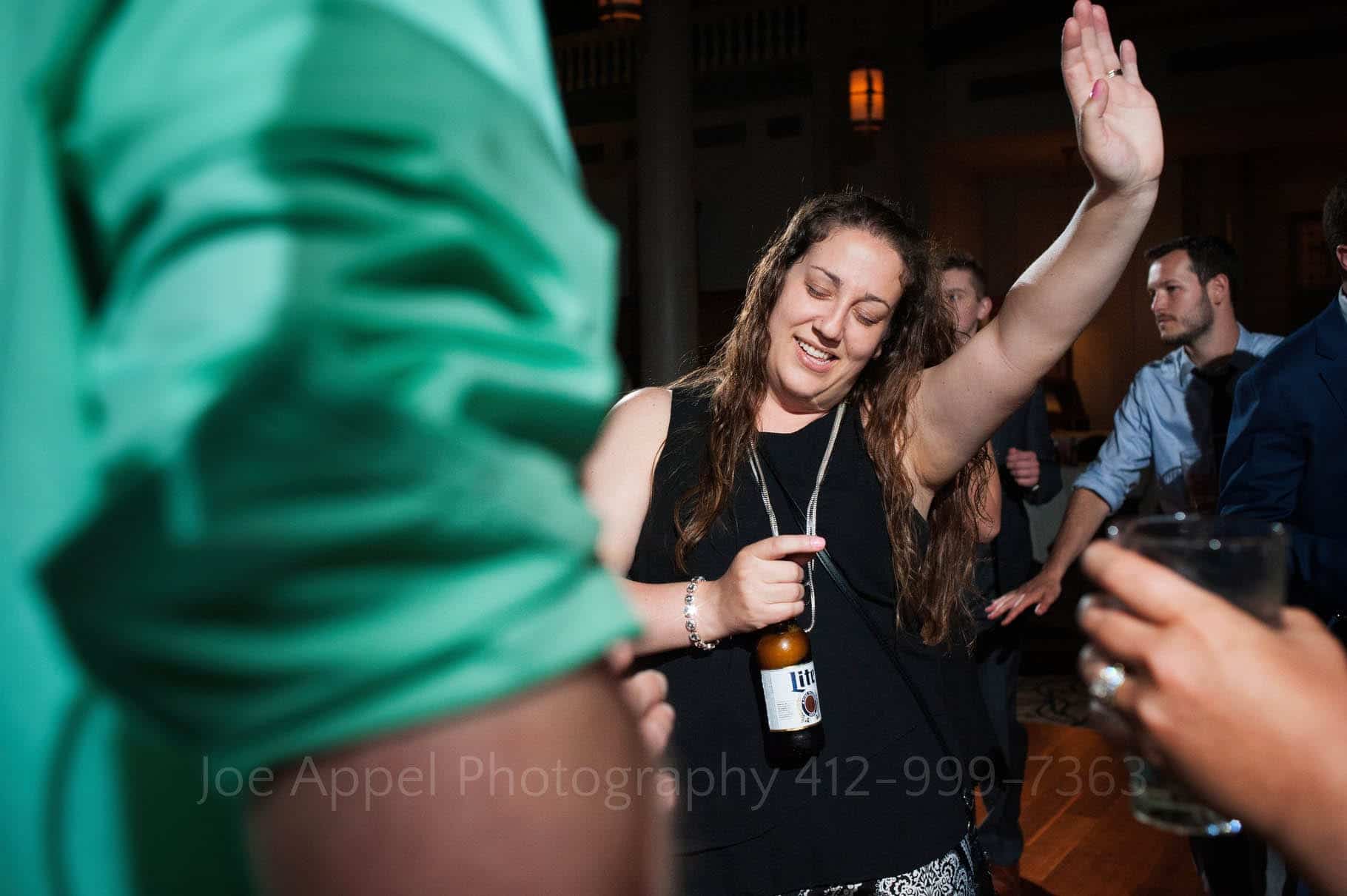 A woman holding a bottle of beer raises her left hand into the air as she dances in front of a man wearing a green shirt.