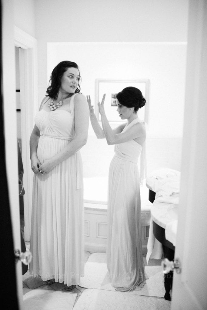 Two bridesmaids stand in a bathroom as they get dressed in their gowns. The taller one looks over her shoulder as the shorter one raises her hands after tying the bow on the back of the first one's dress.