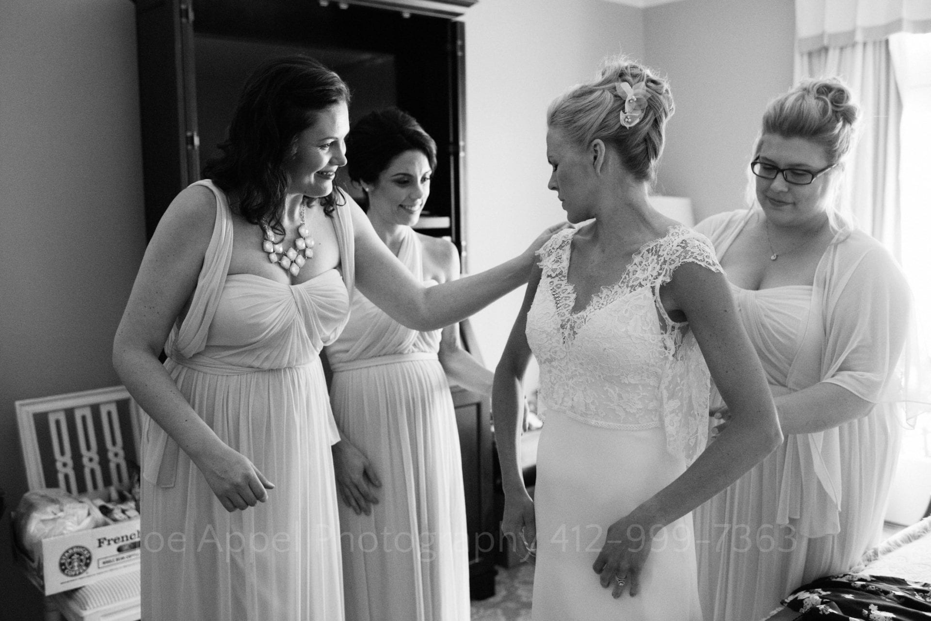 Black and white photo of three bridesmaids helping a bride get her wedding dress on and buttoned up during their Omni Bedford Springs Resort Wedding.