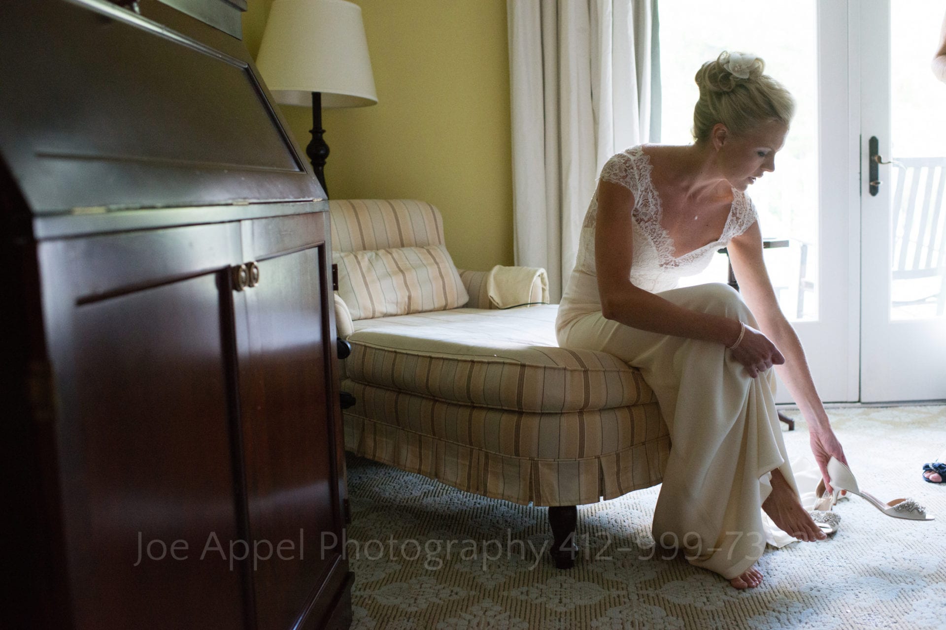 A bride sits on a daybed as she reaches down to put on her open toed shoes during their Omni Bedford Springs Resort Wedding. At the left of the frame is a dark wood cabinet.