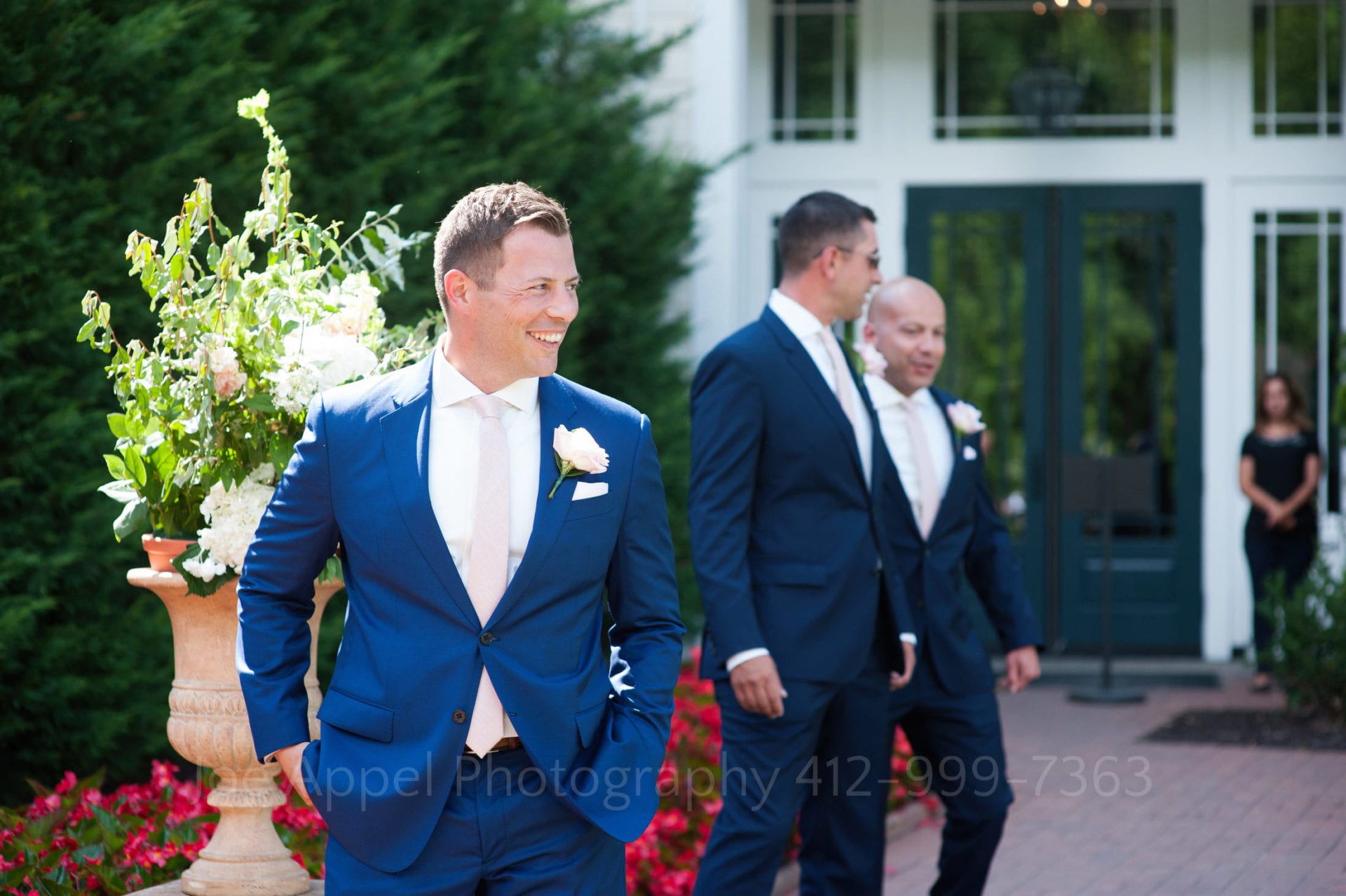 A groom wearing a blue suit and pink tie smiles large as he walks into a courtyard with his hands in his pockets and looks to the right during their Omni Bedford Springs Resort Wedding.
