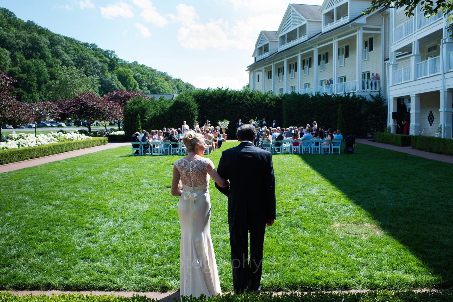 Seen from behind, a bride looks at her father as they stand at the edge of a lawn before they walk to a wedding set up on the other edge of lawn during their Omni Bedford Springs Resort Wedding.