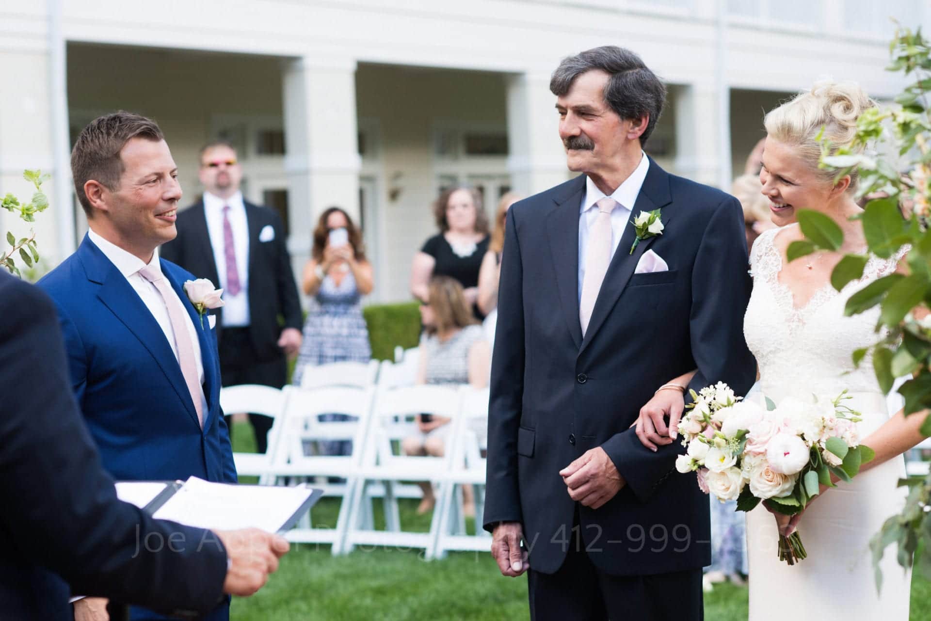 A bride and groom smile at each other as the bride arrives at the end of the aisle with her father during their Omni Bedford Springs Resort Wedding.