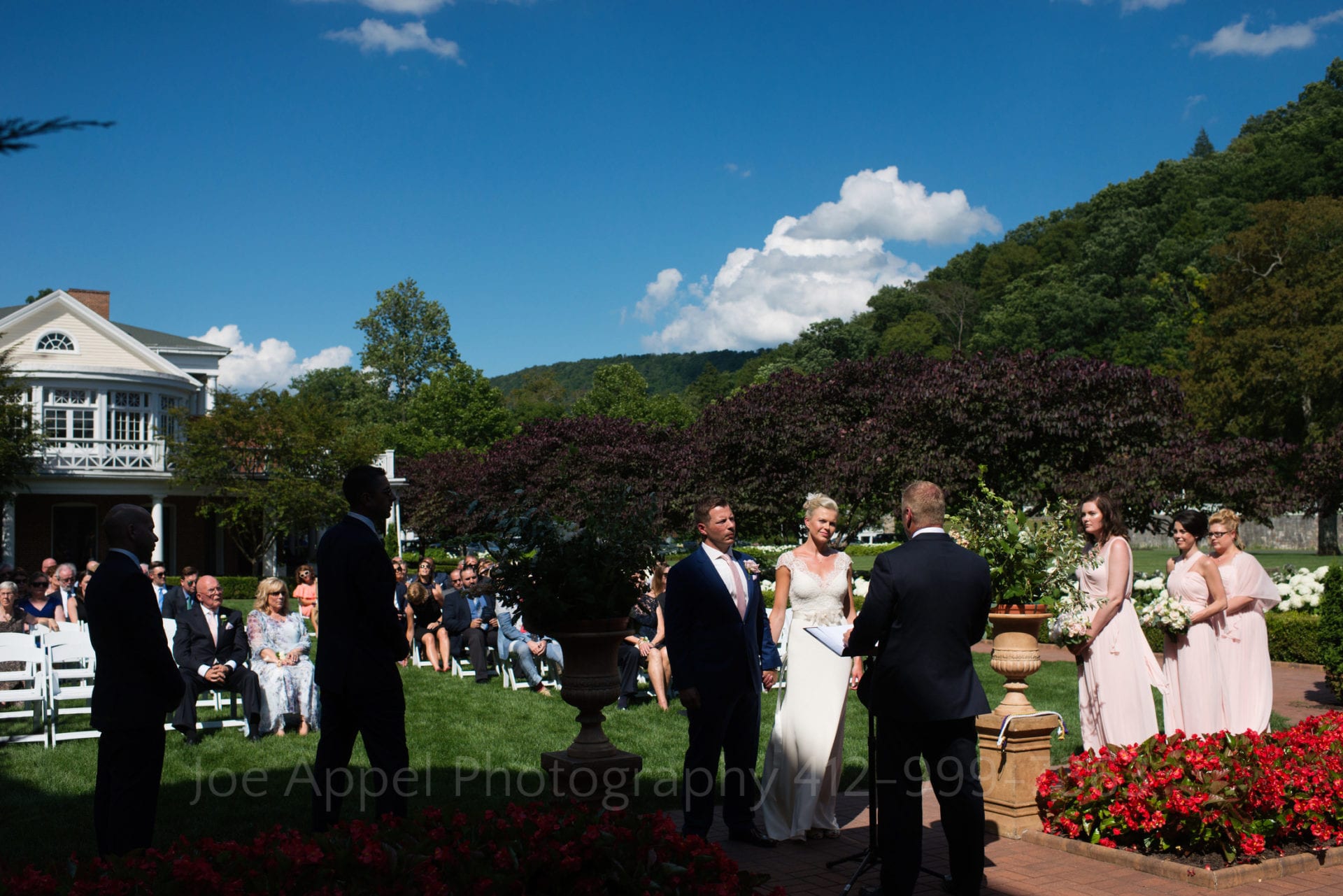With the Allegheny Mountains in the background, a bride and groom stand on a lawn beneath a blue sky during their wedding ceremony during their Omni Bedford Springs Resort Wedding.