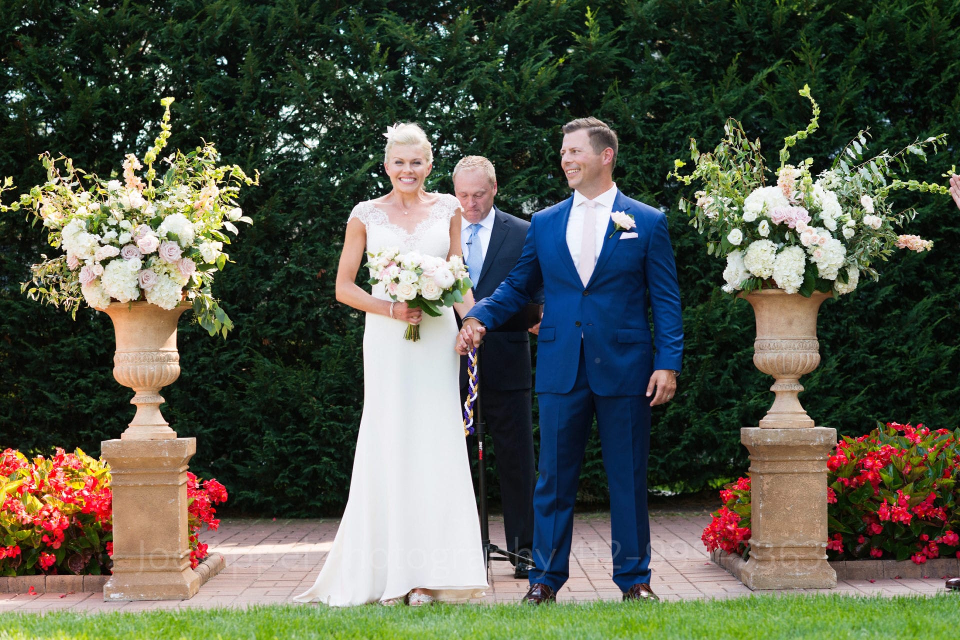 A bride holding a bouquet of roses has a big smile on her face as she and her groom (who looks over at her) are presented to their guests as man and wife. There are two urns filled with flowers and vines to either side of them during their Omni Bedford Springs Resort Wedding.