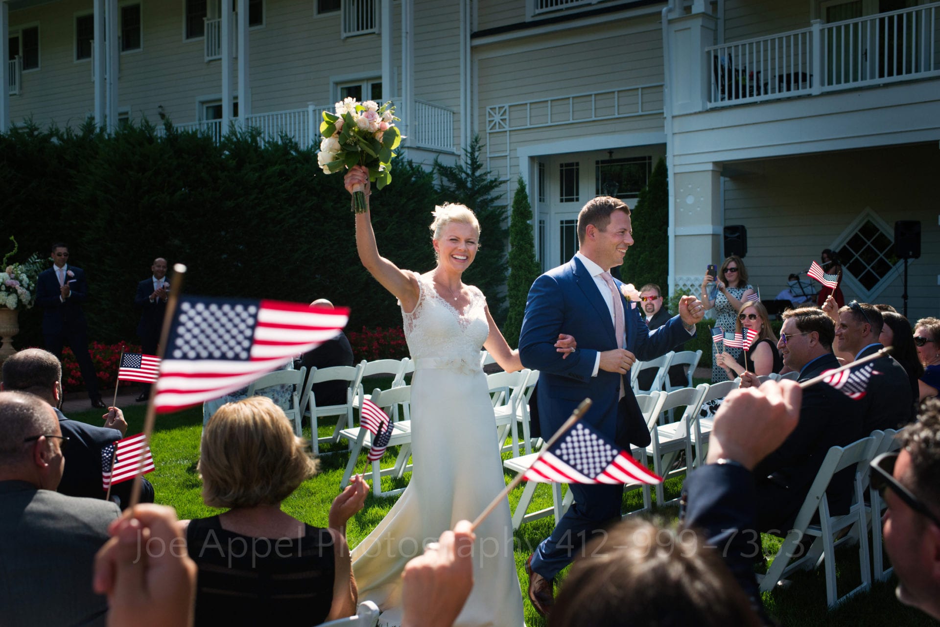 American flags wave as a bride and groom walk down the aisle at the end of their wedding ceremony. The bride raises her bouquet in the air and the groom pumps his fist during their Omni Bedford Springs Resort Wedding.