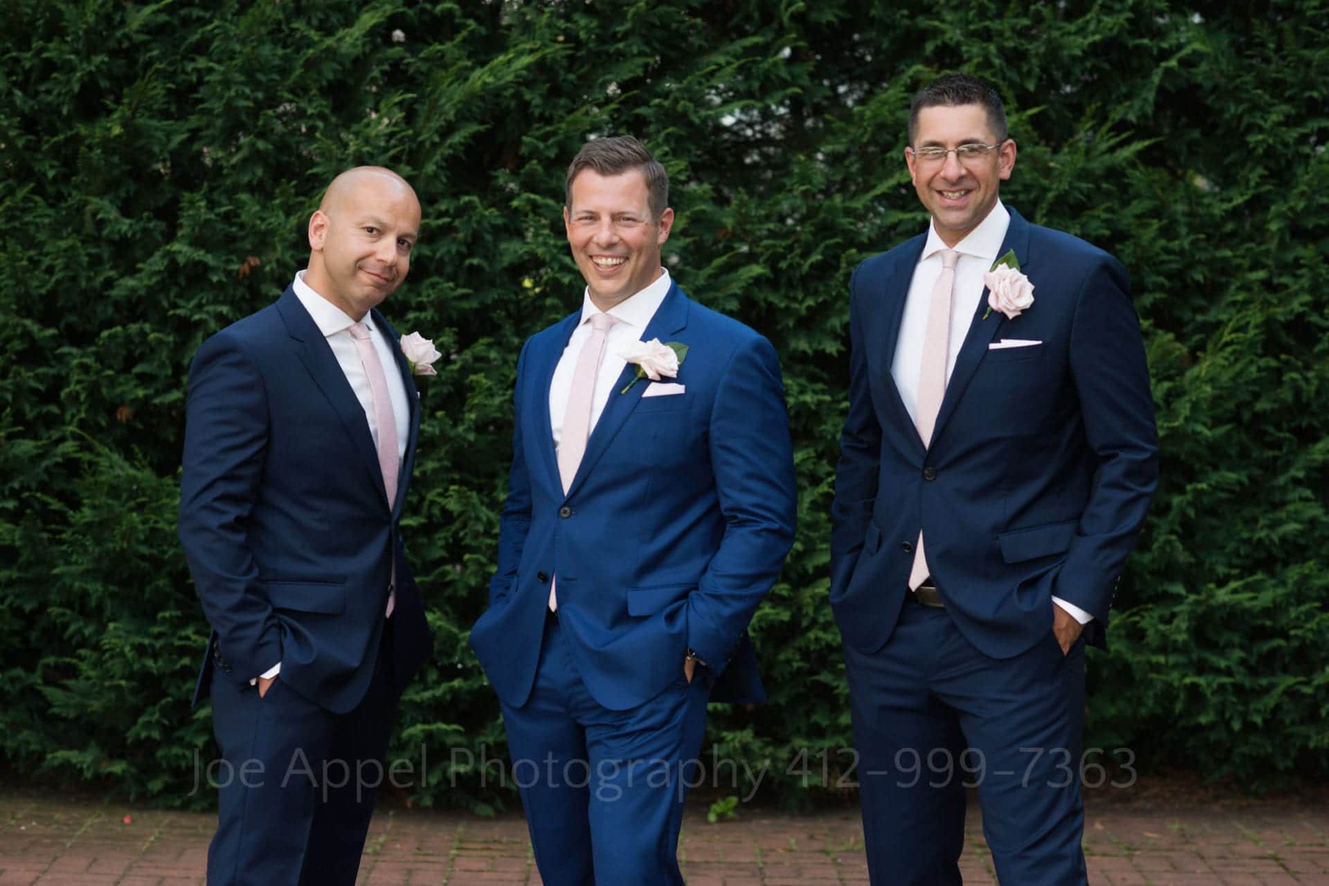 A groom and his two groomsmen stand in their blue suits and pink ties with their hands in their pockets.