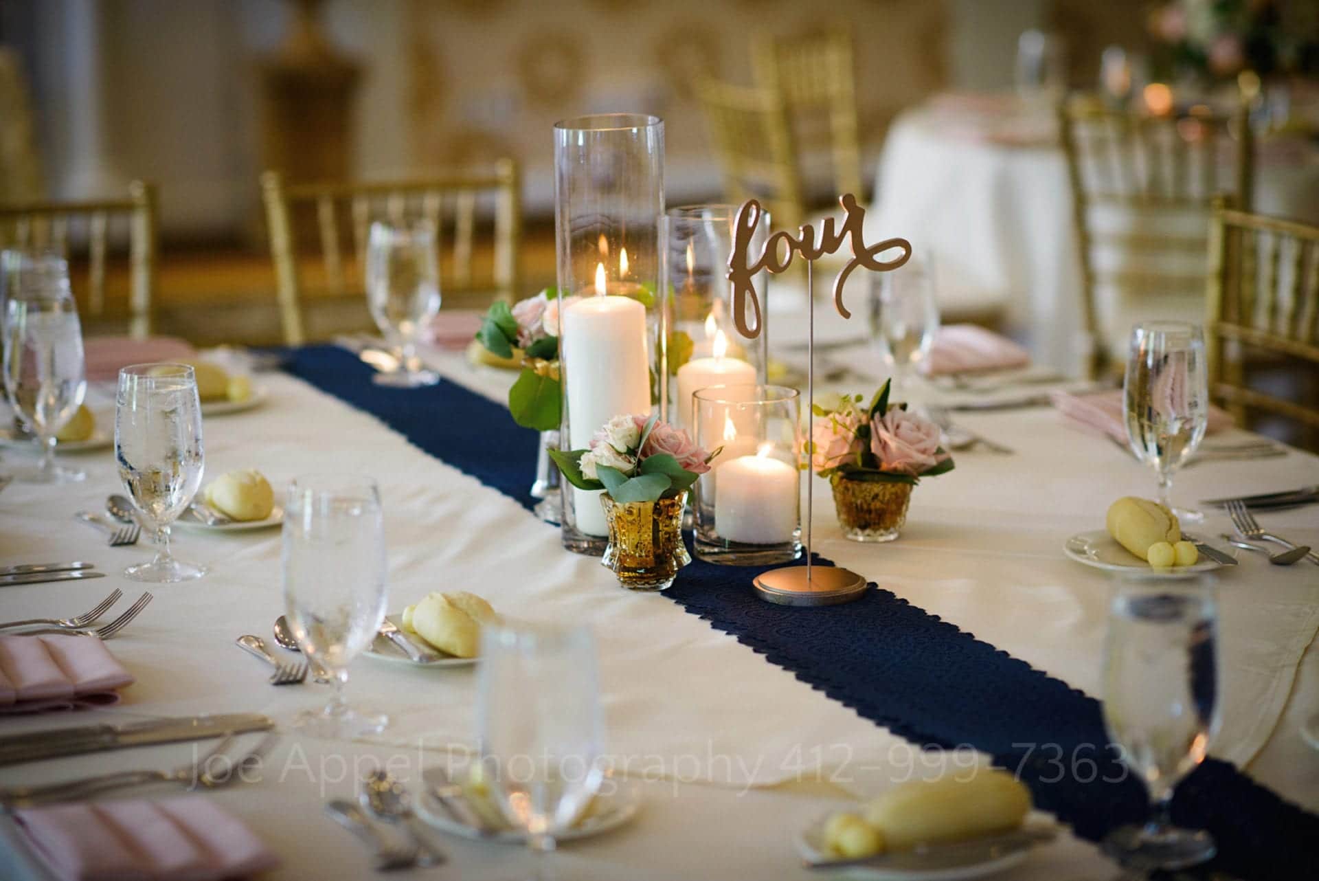 Detail of table four with a centerpiece of candles and small vases of roses. Plates with pink folded napkins and gold chairs. A navy blue runner in the middle during their Omni Bedford Springs Resort Wedding.