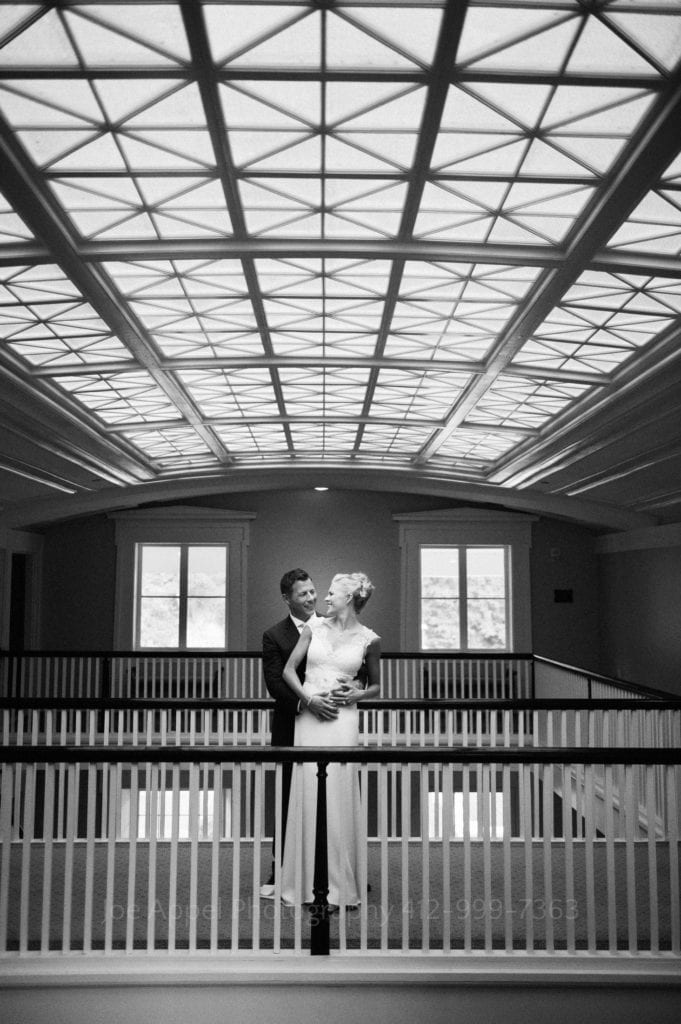 A bride and groom stand together on an elevated walkway in an embrace beneath a large skylight in the Omni Bedford Springs Resort.