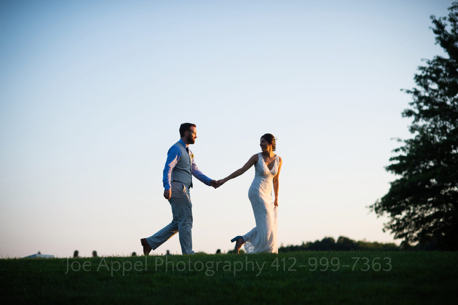 A bride holds hands with her groom while walking in front of him on the top of a hill with a blue sky behind them during their Armstrong Farms Wedding. She looks back at him.