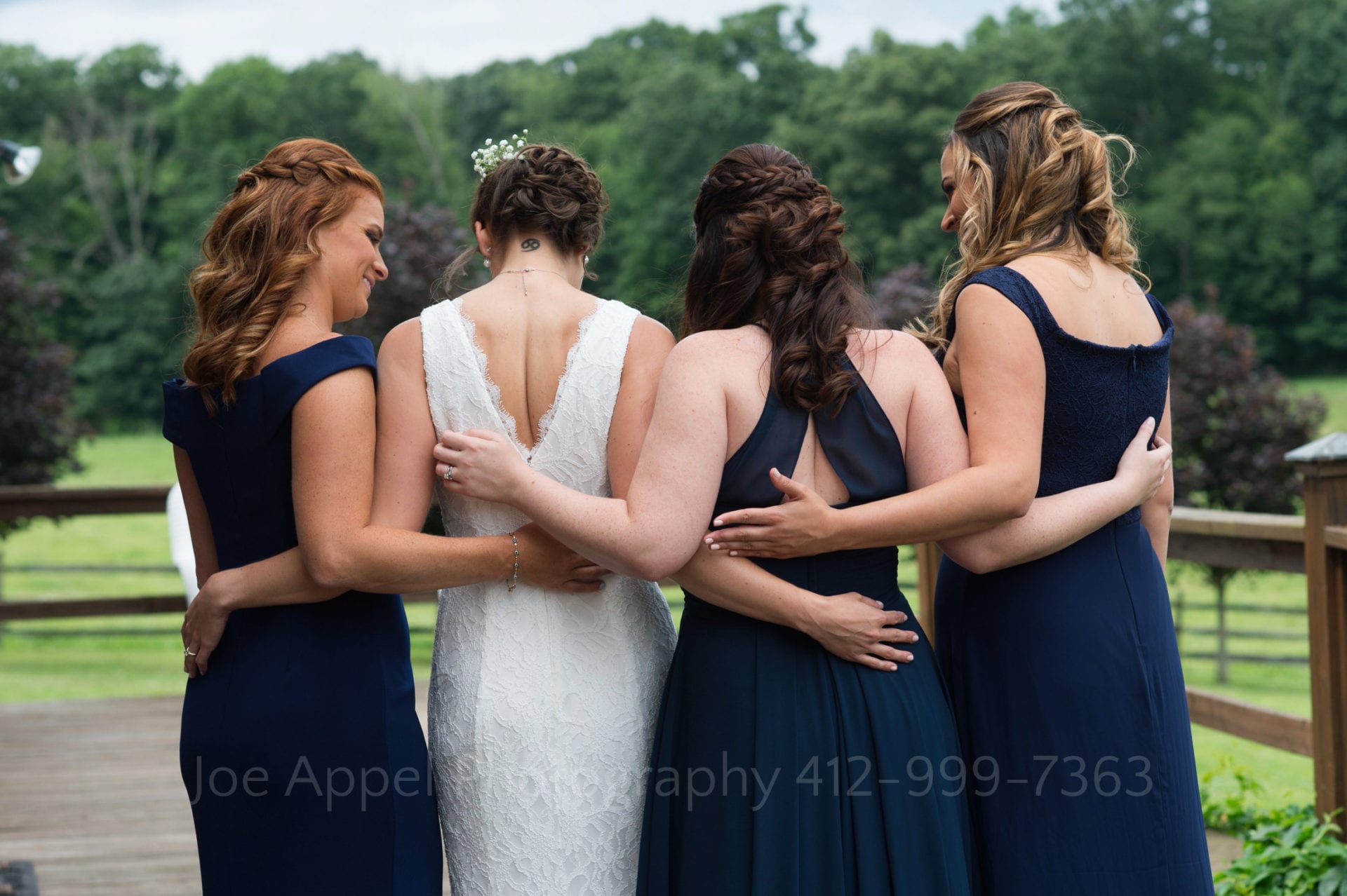 A bride in white and her bridesmaids hold their arms around each other as they stand outside during an Armstrong Farms Wedding. The photo is shot from behind.