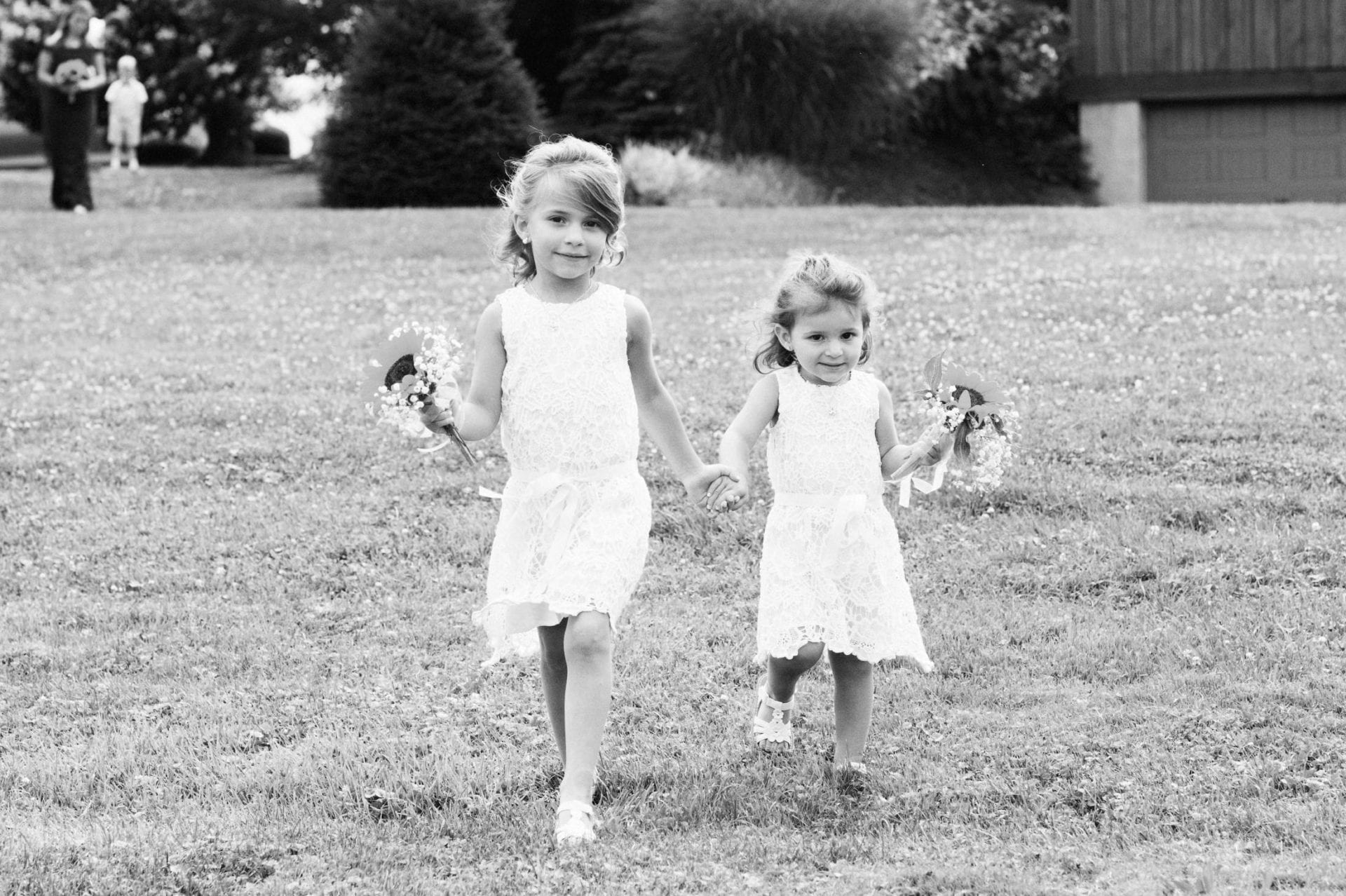 Two young girls wearing white dresses and shoes walk down a hill while carrying flowers during an Armstrong Farms Wedding.