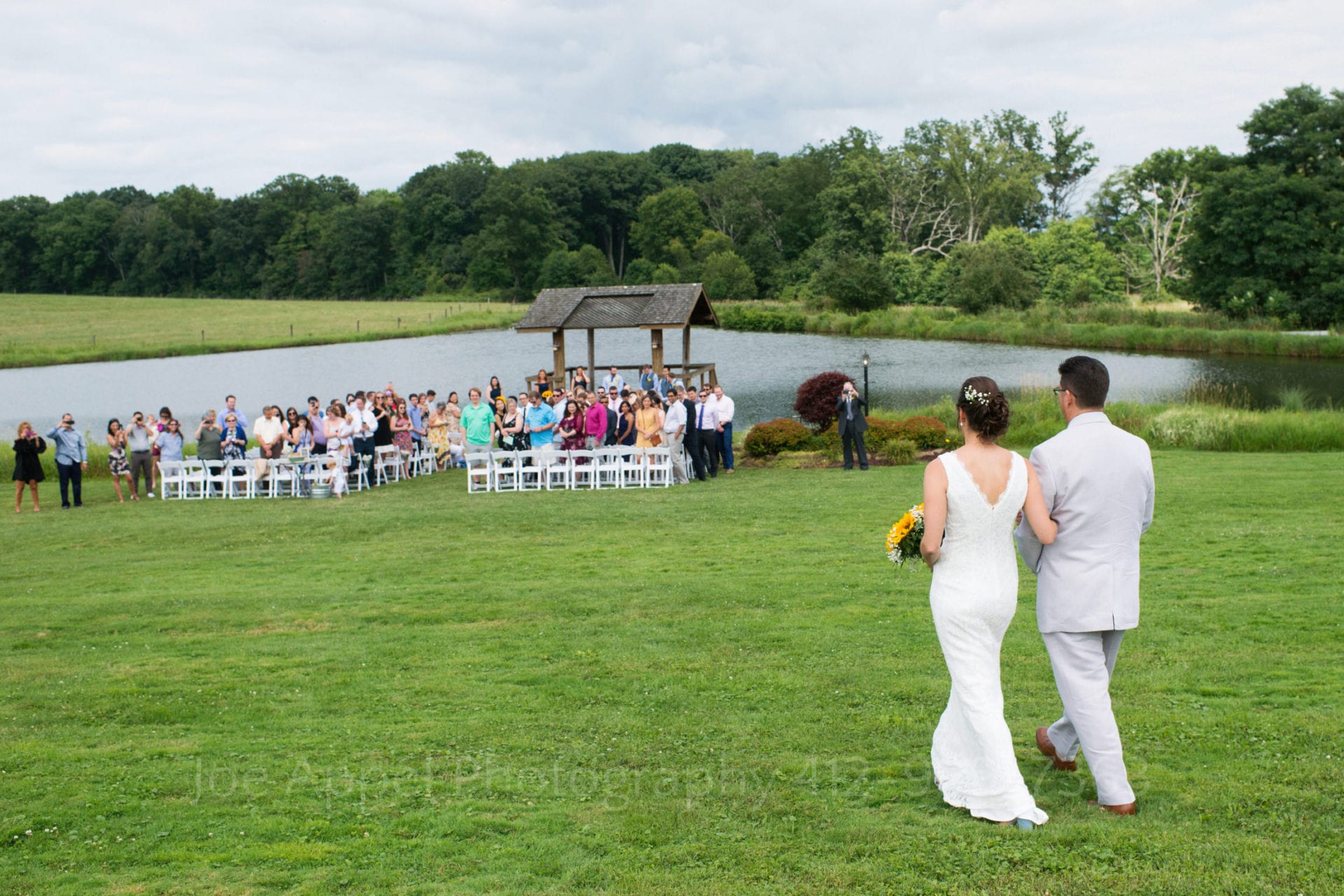 Seen from behind them, a bride and her father walk down a grassy hill towards a pond with a small shelter in front of it. The guests are standing awaiting her arrival to her Armstrong Farms Wedding.