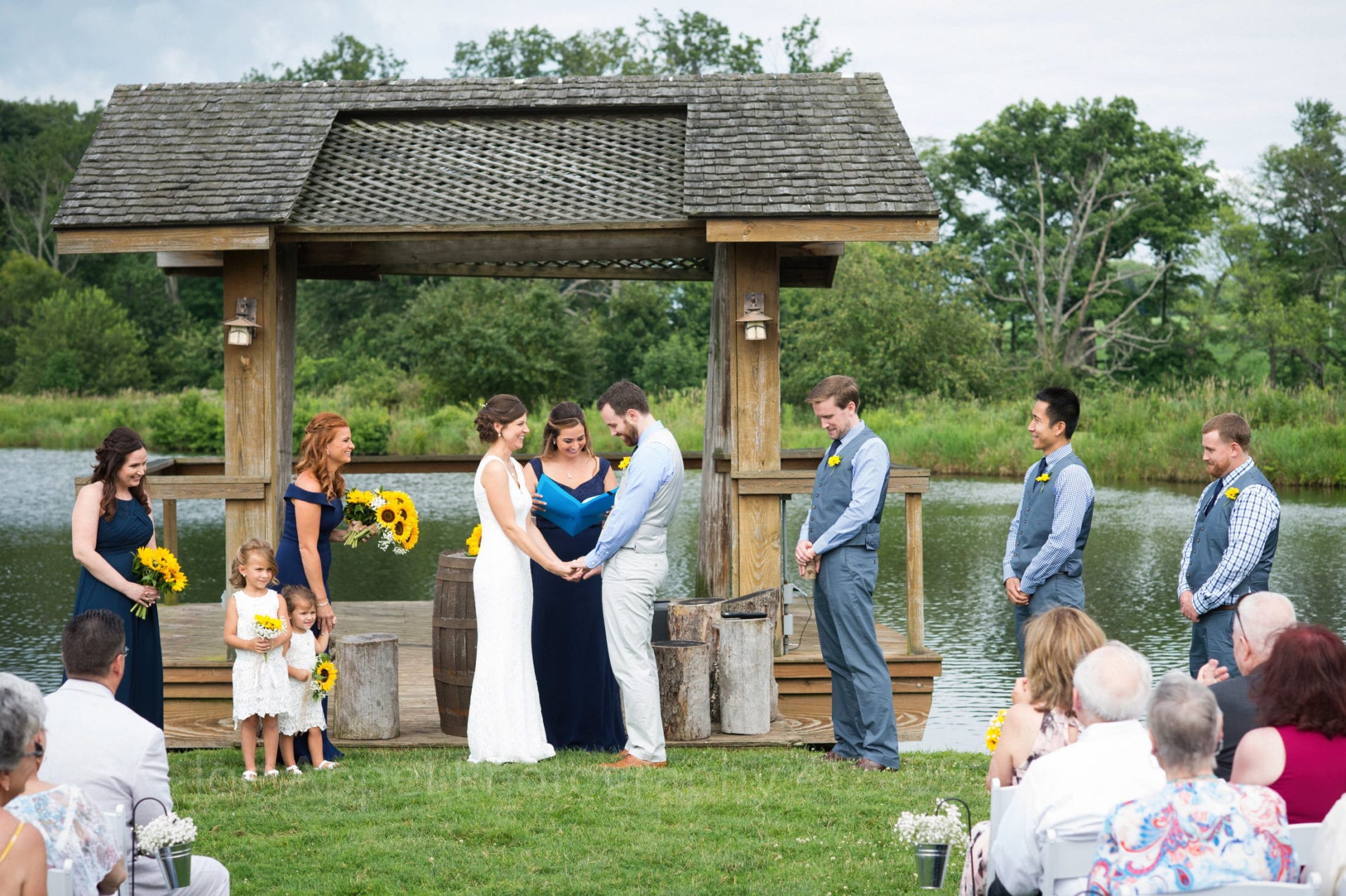 Long shot of a bride and groom exchanging vows in front of a small shelter in a field by a pond at an Armstrong Farms Wedding.