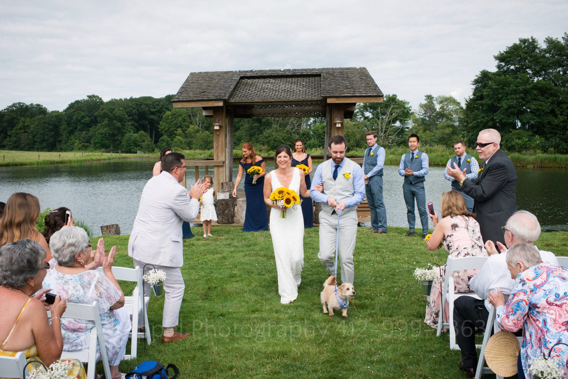 A bride and groom walk down an aisle between chairs set in a field by a pond after their Armstrong Farms Wedding. The groom is holding a leash as their terrier leads them.