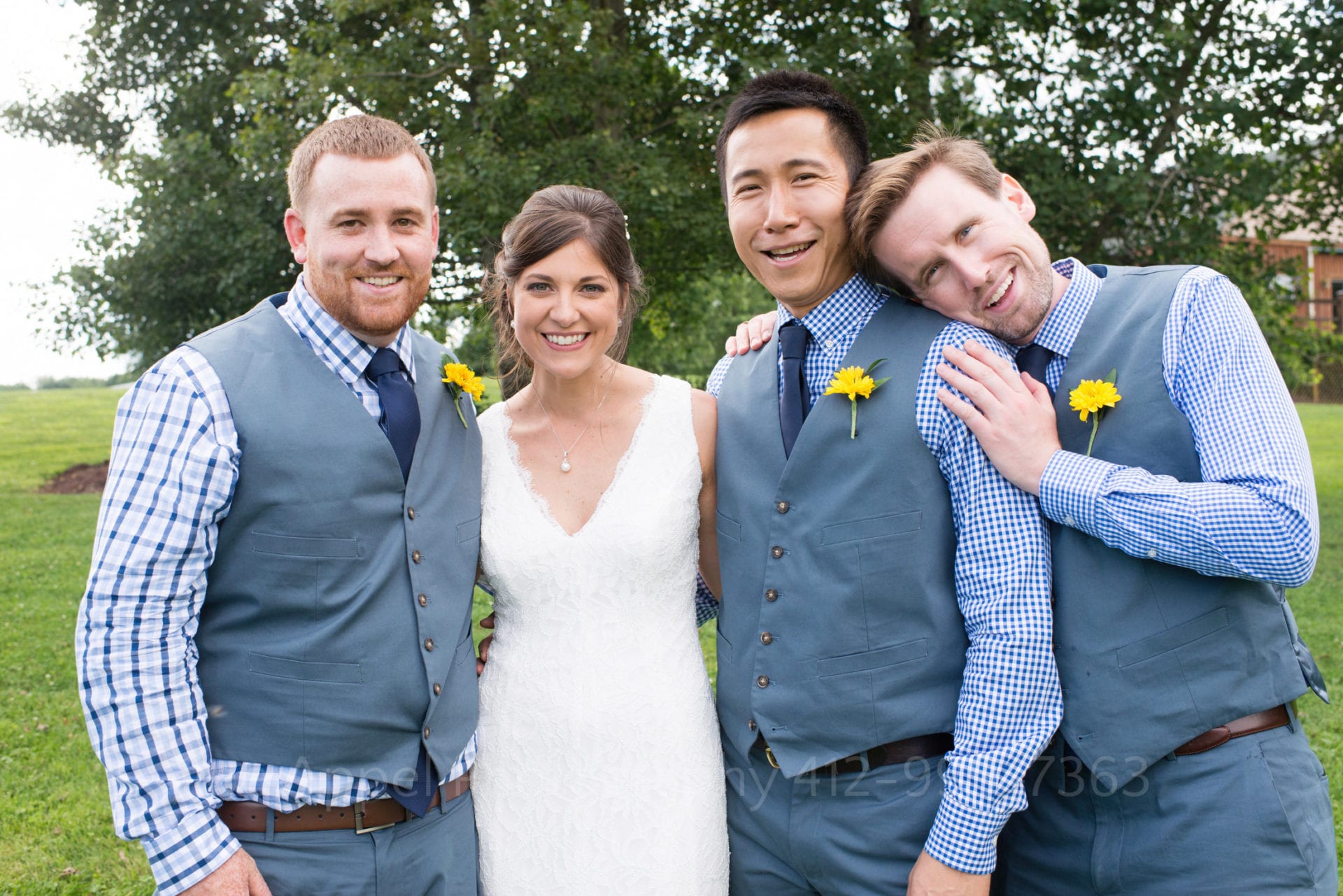 A bride at an Armstrong Farms Wedding smiles as she stands with three groomsmen wearing blue vests and checkered shirts. One groomsman rests his head on the shoulder of another groomsman.