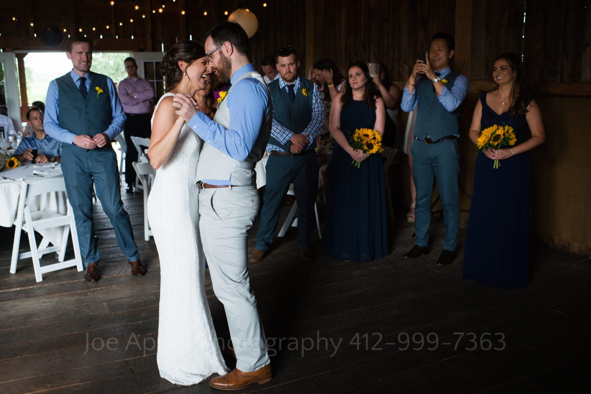 Bride wearing white and groom wearing a tan vest and blue shirt dance close together while their guests surround them.