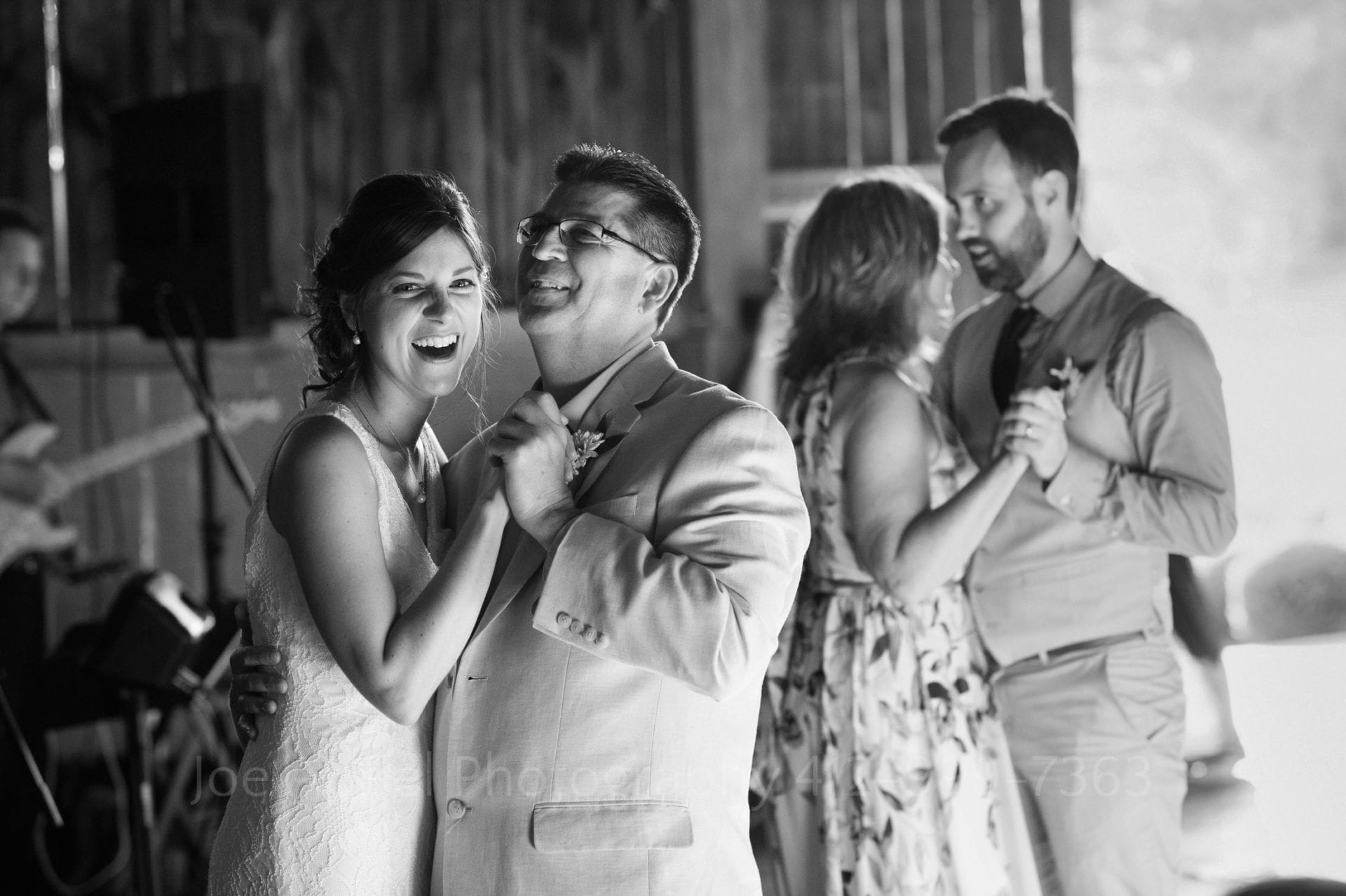 Bride dances with her father - both of them smiling. In the background the groom dances with his mom.