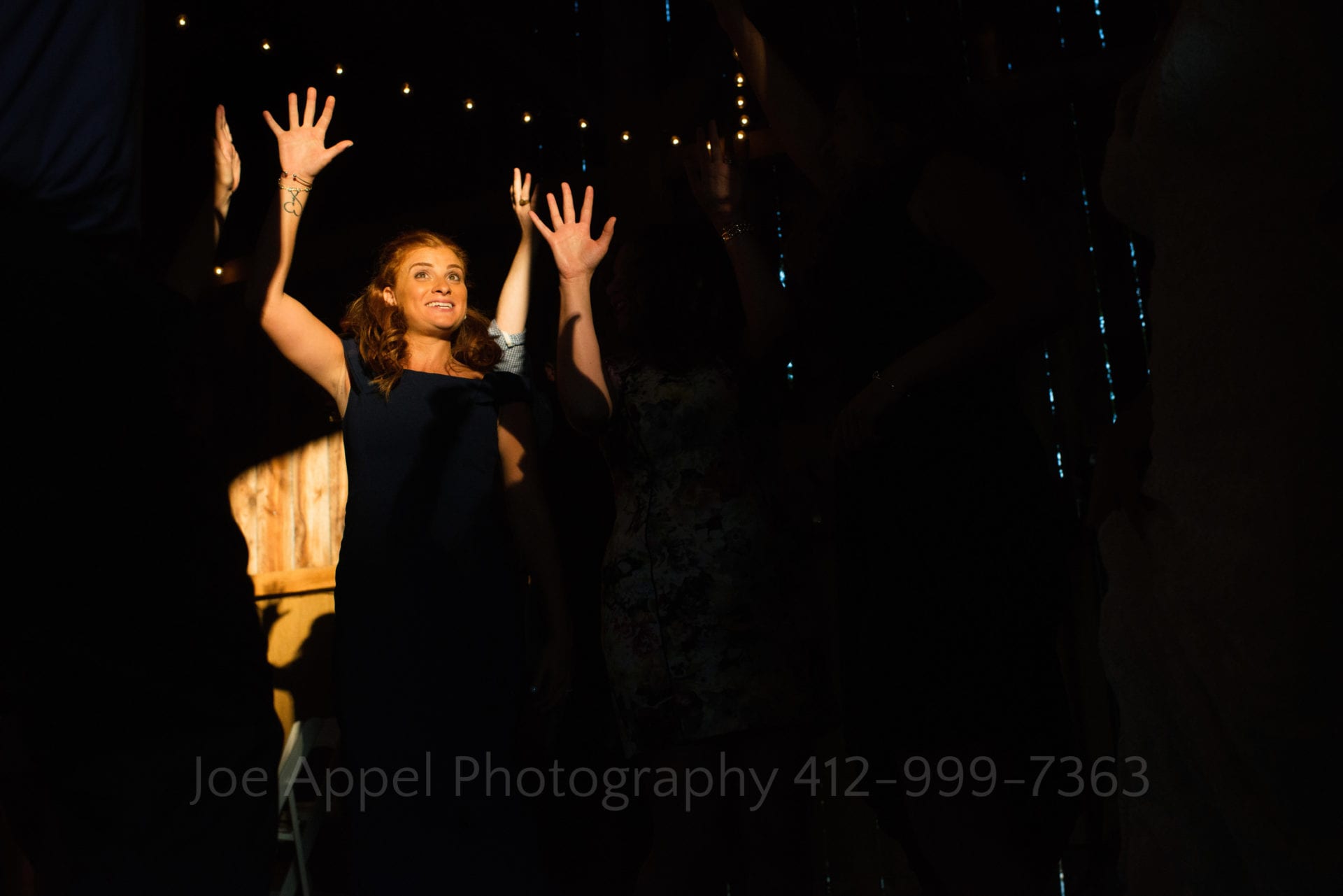 A woman is illuminated by a shaft of golden sunlight while she dances with her hands in the air and her fingers splayed.