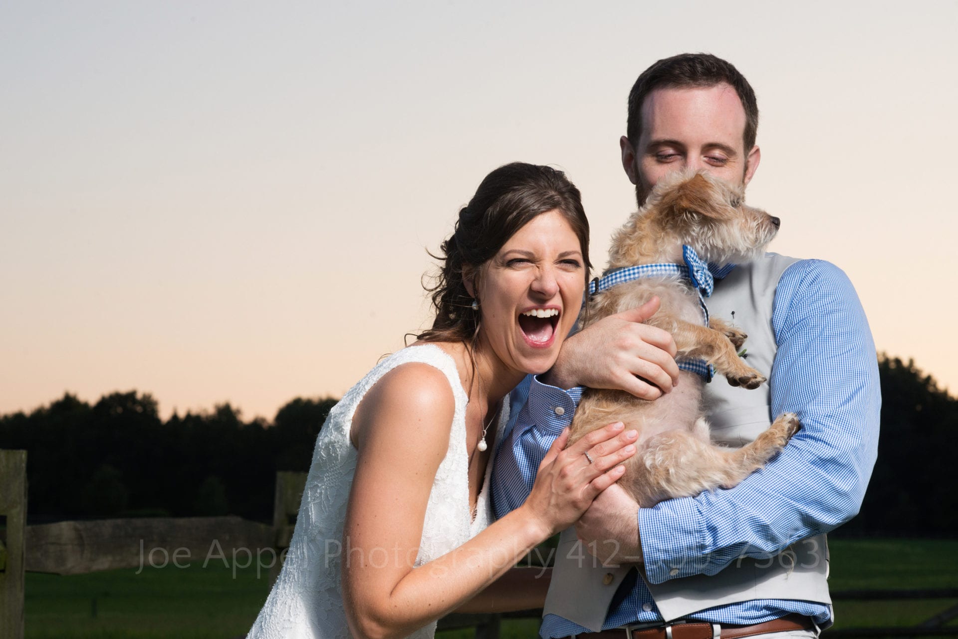 A bride laughs as she and her groom hold their terrier in a family photo.