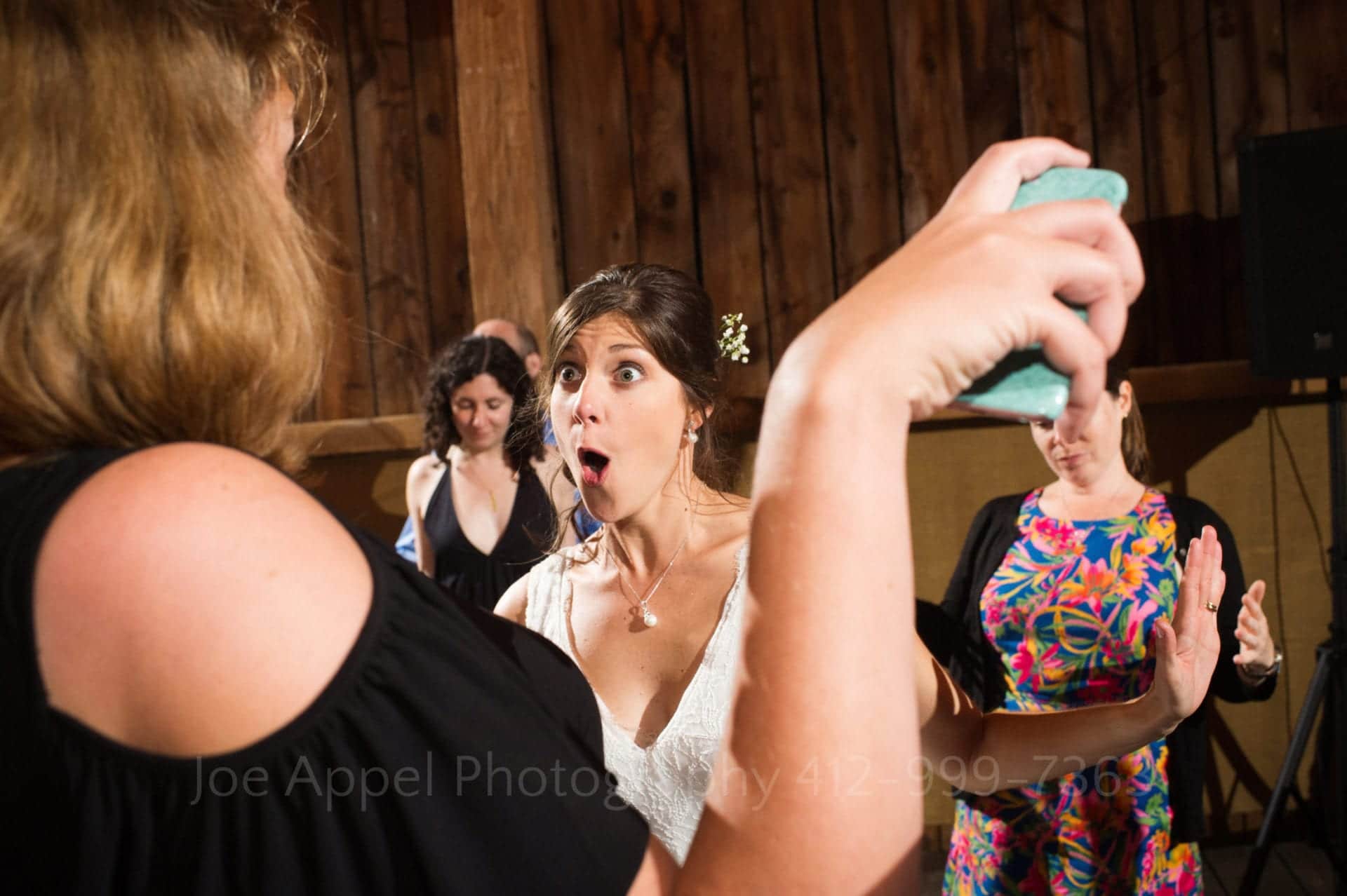 Seen through the outstretched arm of a woman wearing a bare shouldered dress, a bride makes a funny face while dancing.