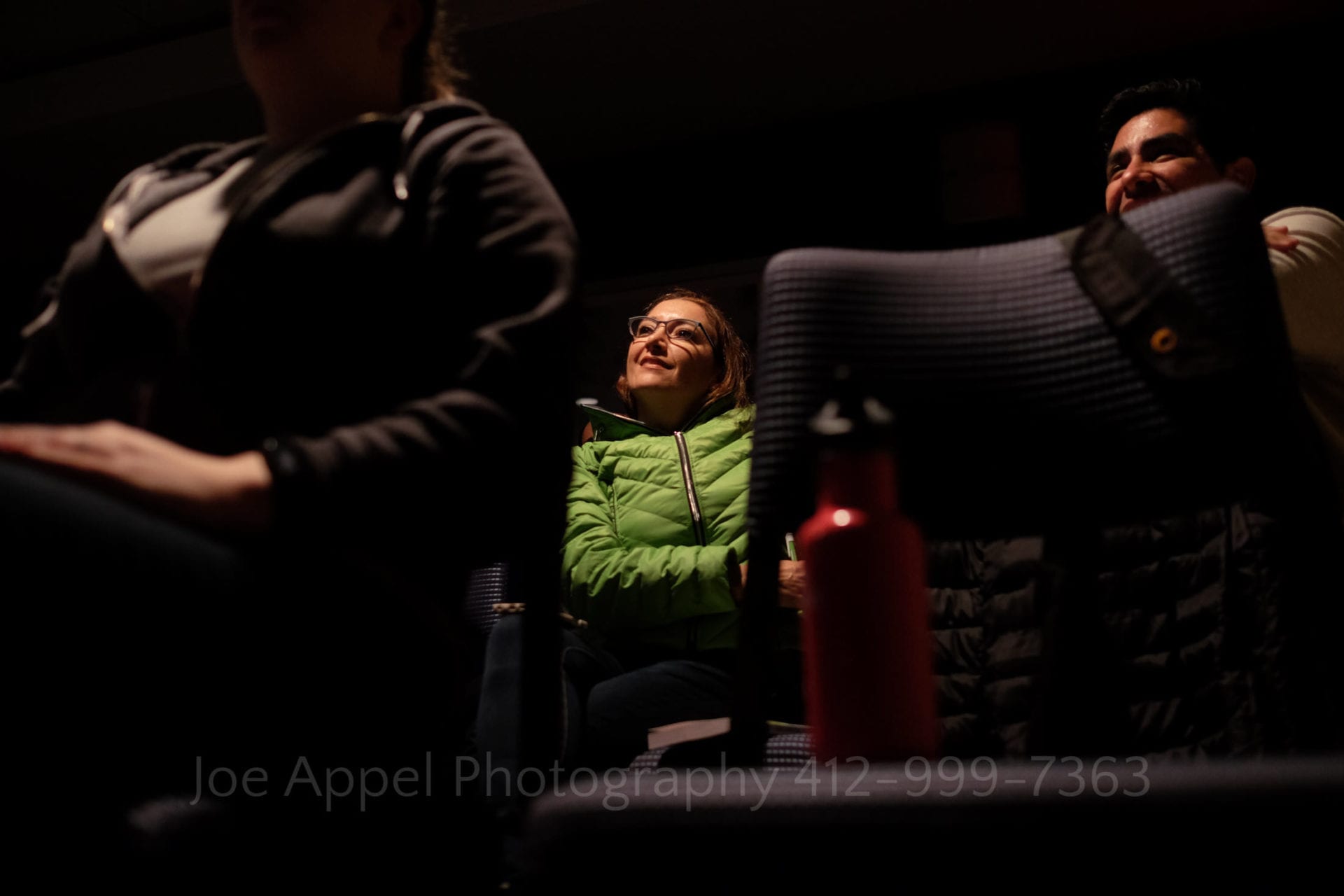 A woman sits in a darkened room while listening to a lecture. she wears a green puffer coat.
